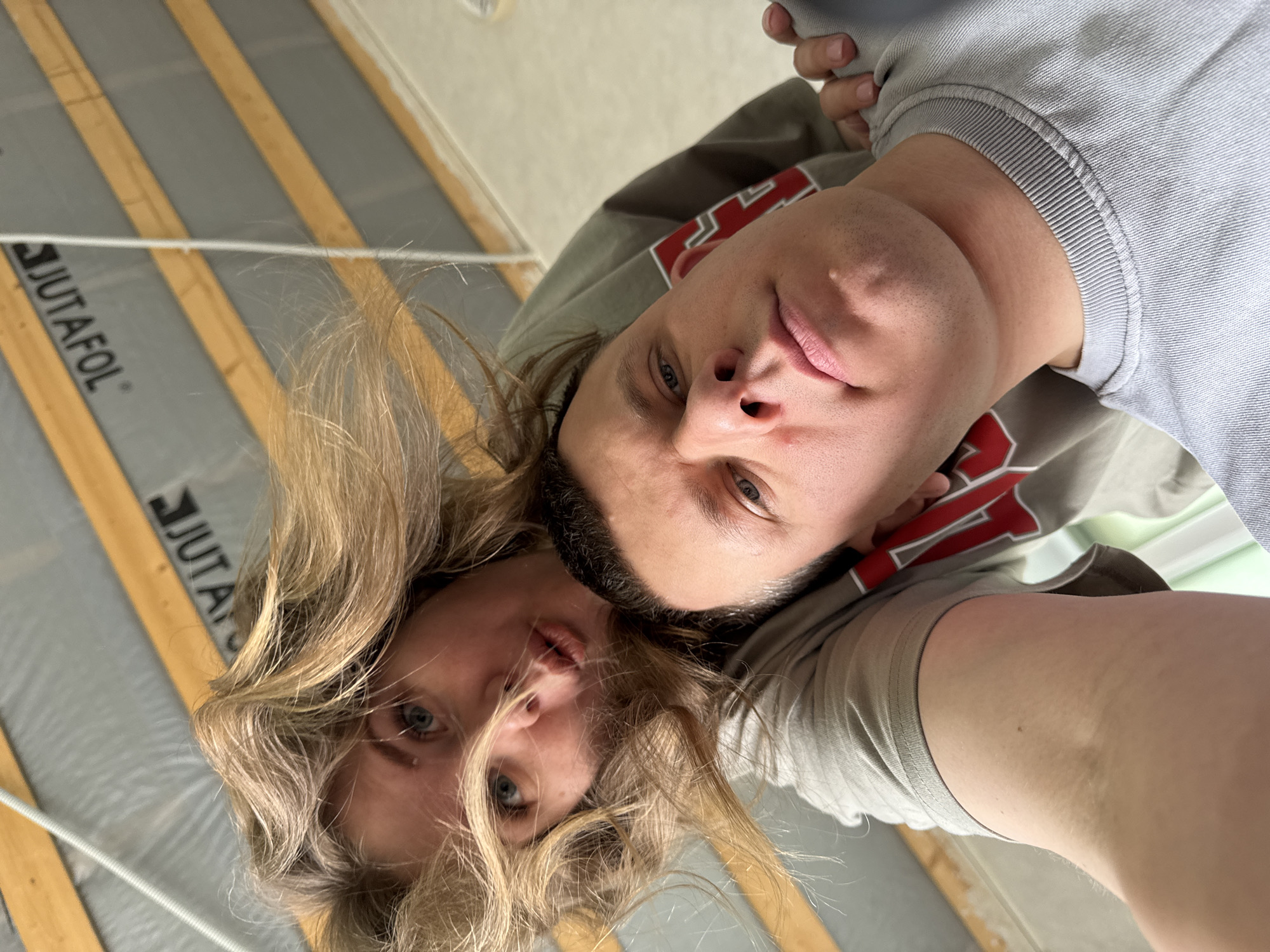 The image shows a close-up, tilted selfie of a woman and a man. The woman has blonde, wavy hair and blue eyes. The man is behind her, wearing a gray sweatshirt. The background reveals unfinished construction, with wooden beams and grey insulation bearing the inscription "JUTAFOL". The location appears to be indoors, possibly in a house or building under construction. While the location is specified as Gornyi (Dvurechenskoe S/P), Russia, no location-specific details are immediately apparent in the image.