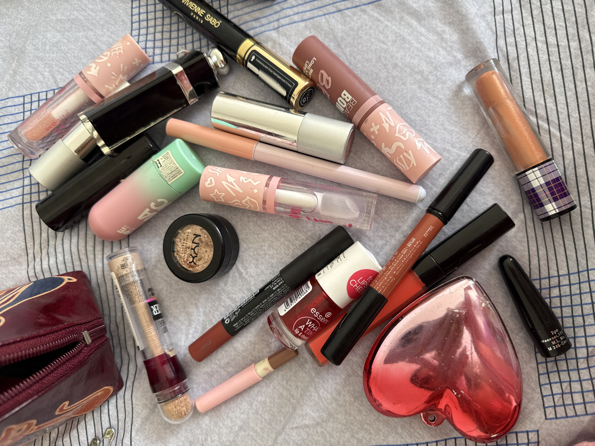 The image shows a collection of makeup products scattered on a striped surface. Items include lipsticks, lip glosses, lip liners, eyeshadow, mascara (Vivienne Sabo), nail polish (Essence What a Tint lip & cheek tint), and a makeup bag. One item mentions "105 USA - 82535 Made in USA" and "Manesar. 122" on a black eyeliner, suggesting the items are from different locations. The city of Moscow, Russia, is not directly indicated in the image.