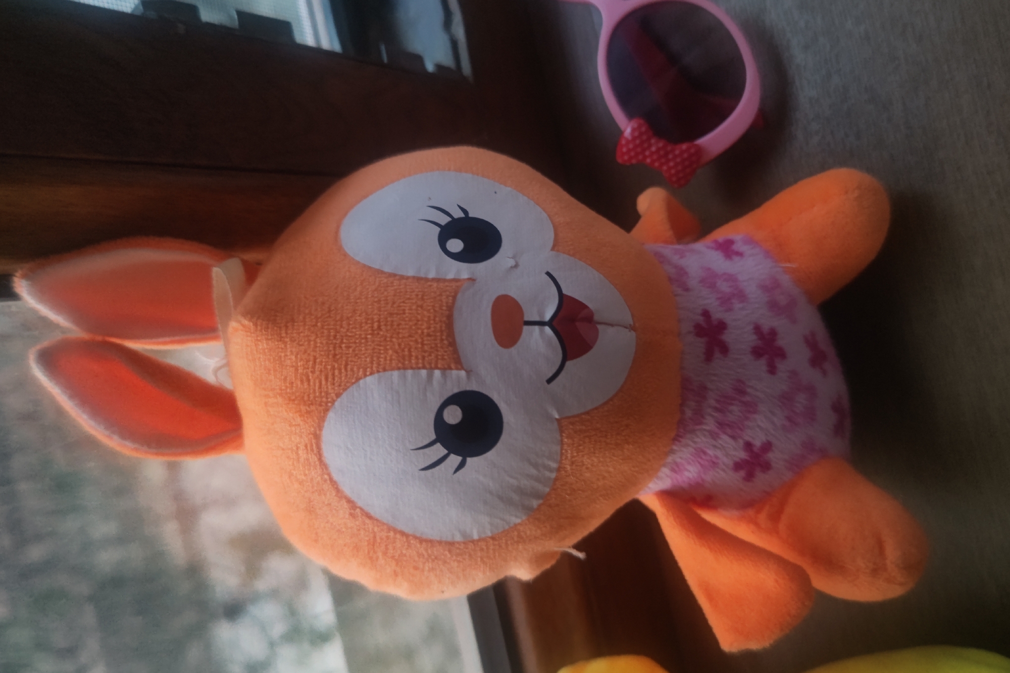 An orange plush bunny toy with large white eyes, pink inside ears, and a pink floral patterned bodysuit sits on a surface next to a pair of pink sunglasses with a red bow. The bunny is facing forward, with its arms slightly spread. The background appears to be a window with blinds and a wooden frame. The scene is indoors and appears to be a close-up shot of the toy. There is no indication of the city of Samarkand, Uzbekistan.