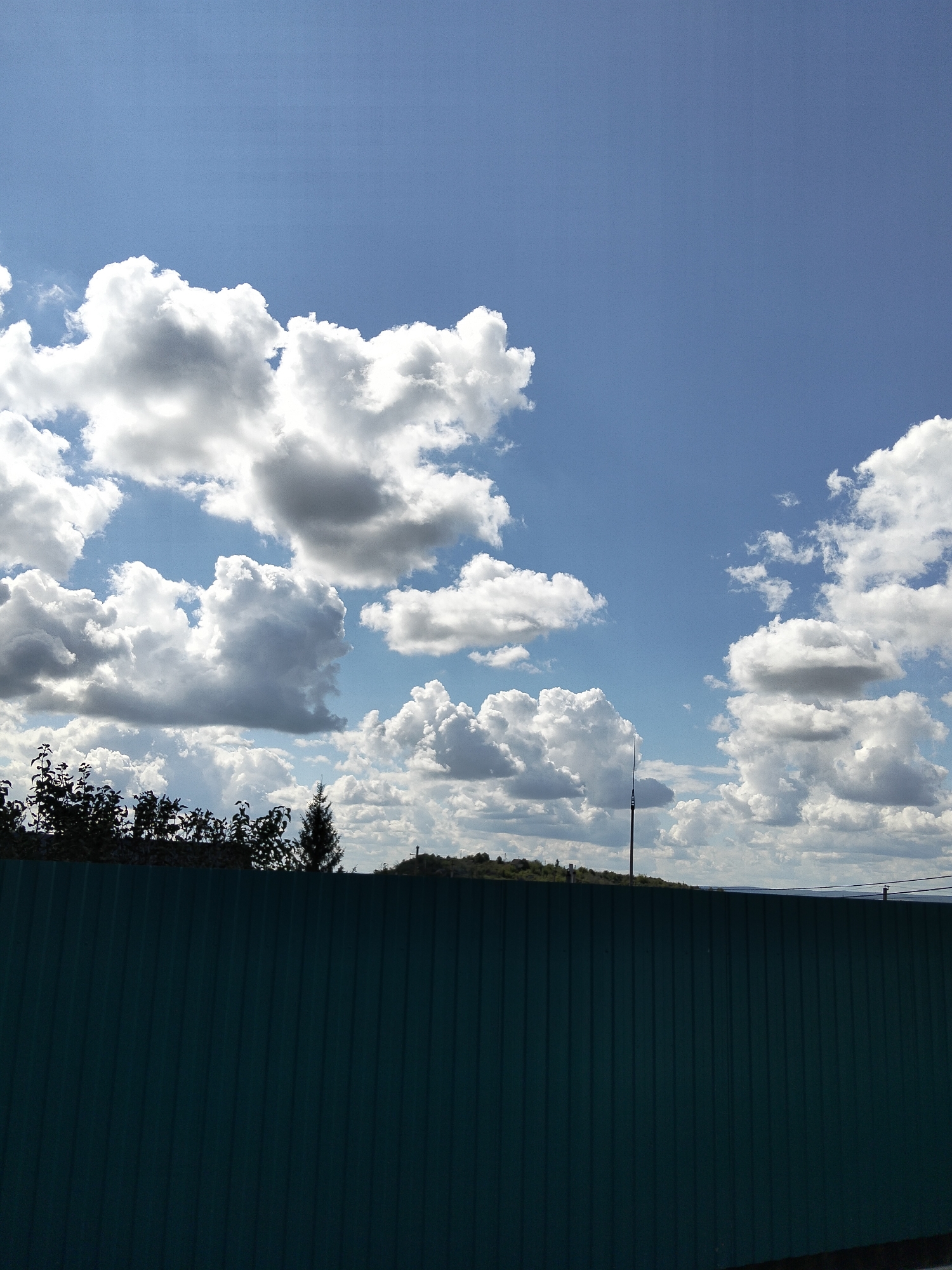 A wide-angle, eye-level, outdoor photograph captures a clear blue sky filled with dramatic, fluffy white and grey clouds. In the foreground, a dark green corrugated metal fence stretches horizontally across the frame, obscuring the lower portion of the scene. Above the fence, a line of dark green trees and foliage is silhouetted against the sky, with a few taller, darker evergreen trees interspersed among them. A slender, dark pole with a pointed top, possibly a lightning rod or antenna, stands vertically in the middle ground. Further in the distance, a low, green, rolling hill or embankment is visible, with hints of structures and a distant body of water suggesting a landscape outside of an urban core, possibly the outskirts of Oktyabrsky, Russia, given the context. The overall lighting suggests daytime, likely a sunny afternoon with scattered clouds. There are no people or visible animals in the image.