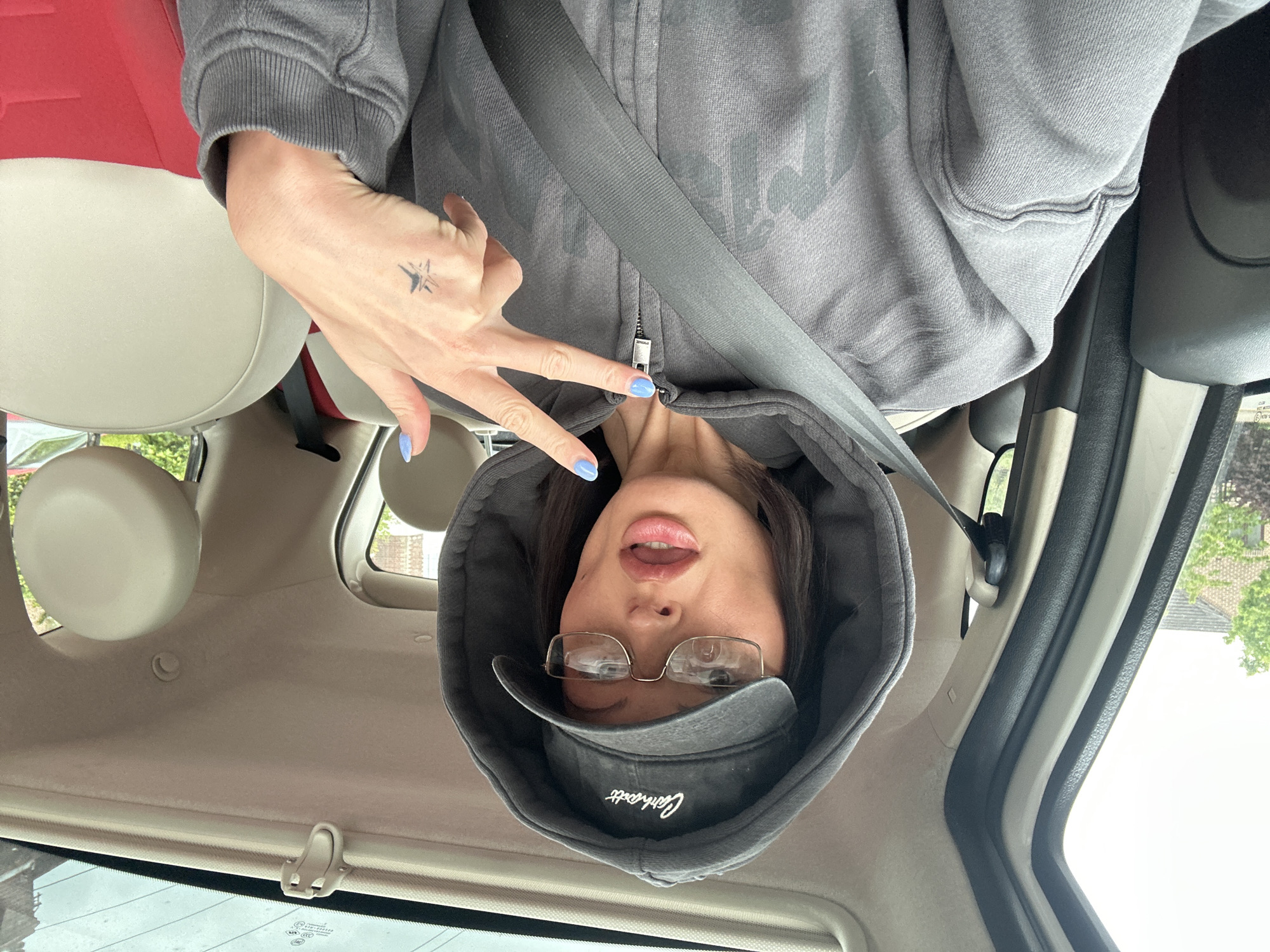 A woman wearing a gray hooded sweatshirt, glasses, and a Carhartt hat is in the driver's seat of a car. The image is oriented upside down. She has blue nail polish and a tattoo on her hand. She is wearing a seatbelt. The interior of the car has white or beige seats and trim, with a red headrest visible. The car is likely in Tunbridge Wells, United Kingdom. Evidence for this is in the car's registration in the window.