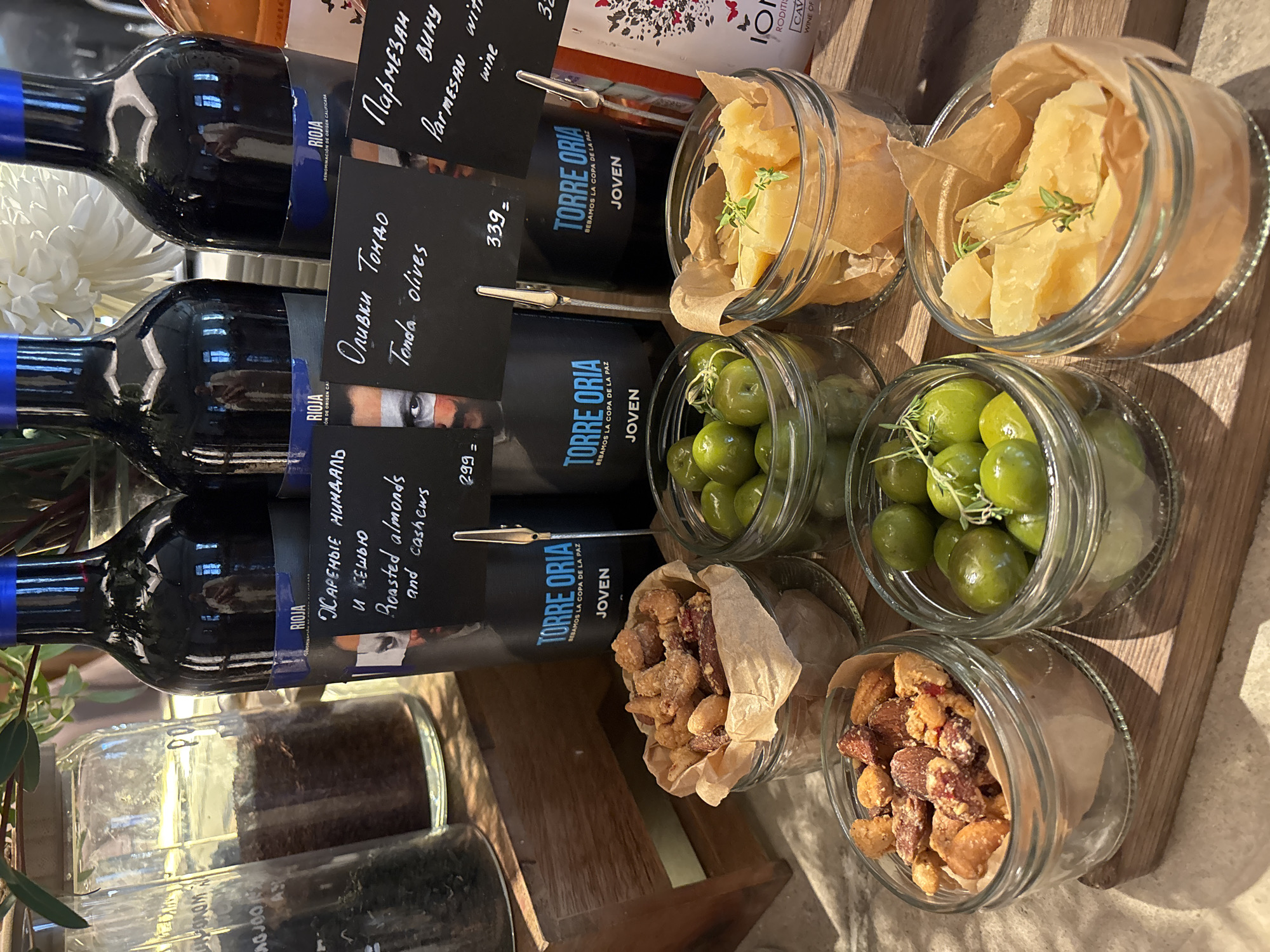 The image displays a close-up of a wooden serving tray at a food establishment in Saint Petersburg, Russia. The tray holds several glass jars filled with food items, likely tapas or appetizers, accompanied by bottles of wine and a tall glass jar containing a dark substance, possibly coffee beans.
In the foreground, three glass jars are arranged. The leftmost jar contains a mixture of nuts, including almonds and cashews, coated in a glaze. The middle jar is filled with green olives, some of which have sprigs of rosemary. The rightmost jar holds chunks of aged cheese, garnished with a sprig of thyme.
Behind these jars, three dark glass bottles of wine are prominently displayed. Each bottle has a label indicating "RIOJA JOVEN" and features a small black sign clipped to its neck. These signs bear descriptions of the accompanying food items in both Russian and English. The Russian text on the first sign reads "ЖАРЕНЫЕ МИНДАЛЬ И КЕШЬЮ," translating to "Roasted almonds and cashews." The second sign reads "ОЛИВКИ ТОНА" in Russian and "Tonda olives" in English. The third sign has "ПАРМЕЗАН ВИНУ" in Russian and "Parmesan wine" in English. The prices "299 =" and "339 =" are visible on two of these signs.
To the left of the wine bottles, a tall, clear glass jar contains a dark, granular substance, likely ground coffee. Next to it, another glass jar, partially obscured, also appears to contain a dark, dry ingredient.
The background features a blurred white floral arrangement, possibly a chrysanthemum, and a glimpse of green foliage, suggesting a decorative element within the establishment. The overall setting appears to be a cafe or restaurant specializing in Spanish-inspired cuisine or wine. The lighting is soft and natural, highlighting the textures and colors of the food and drinks. The image is composed to emphasize the artisanal presentation of the food items.