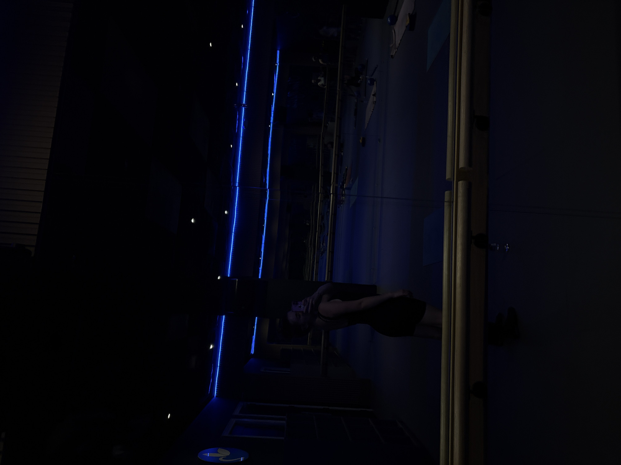 A vertical, low-light interior shot depicts a room with various exercise participants. At least one person is positioned closest to the viewer, while others are faintly visible in the deeper background of the photo. The room is illuminated with blue lights. The scene is possibly in Mexico City, Mexico.