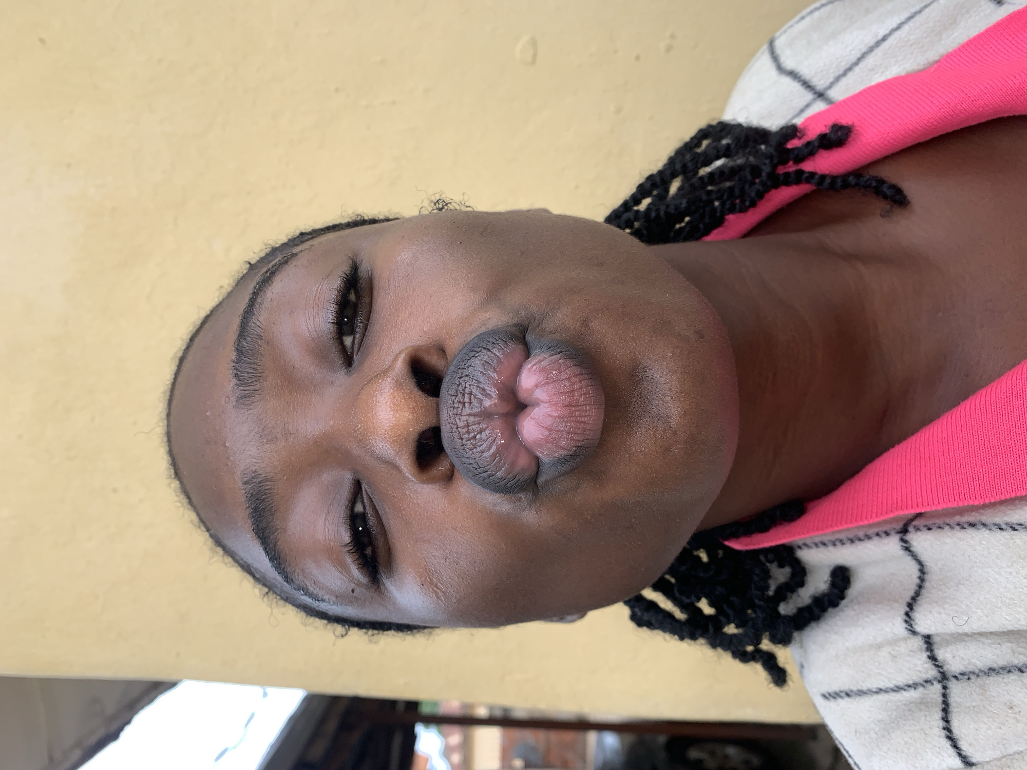 A headshot-style close-up portrait depicts an adult female with dark skin, located in Rafin Par, Nigeria. She faces the camera directly, her eyes open and looking forward, and her lips are prominently puckered in a "kissing" expression. Her dark hair is styled in numerous small braids, visible framing her face and shoulders. She wears a bright fuchsia-pink garment beneath an off-white jacket or cardigan with a black grid pattern. The background on the right and top is a textured, light yellow wall. To the far left, blurred architectural elements are visible, including a light-colored structure in the upper corner and indistinct outdoor features in the lower left, possibly suggesting a street or partial view of a vehicle.