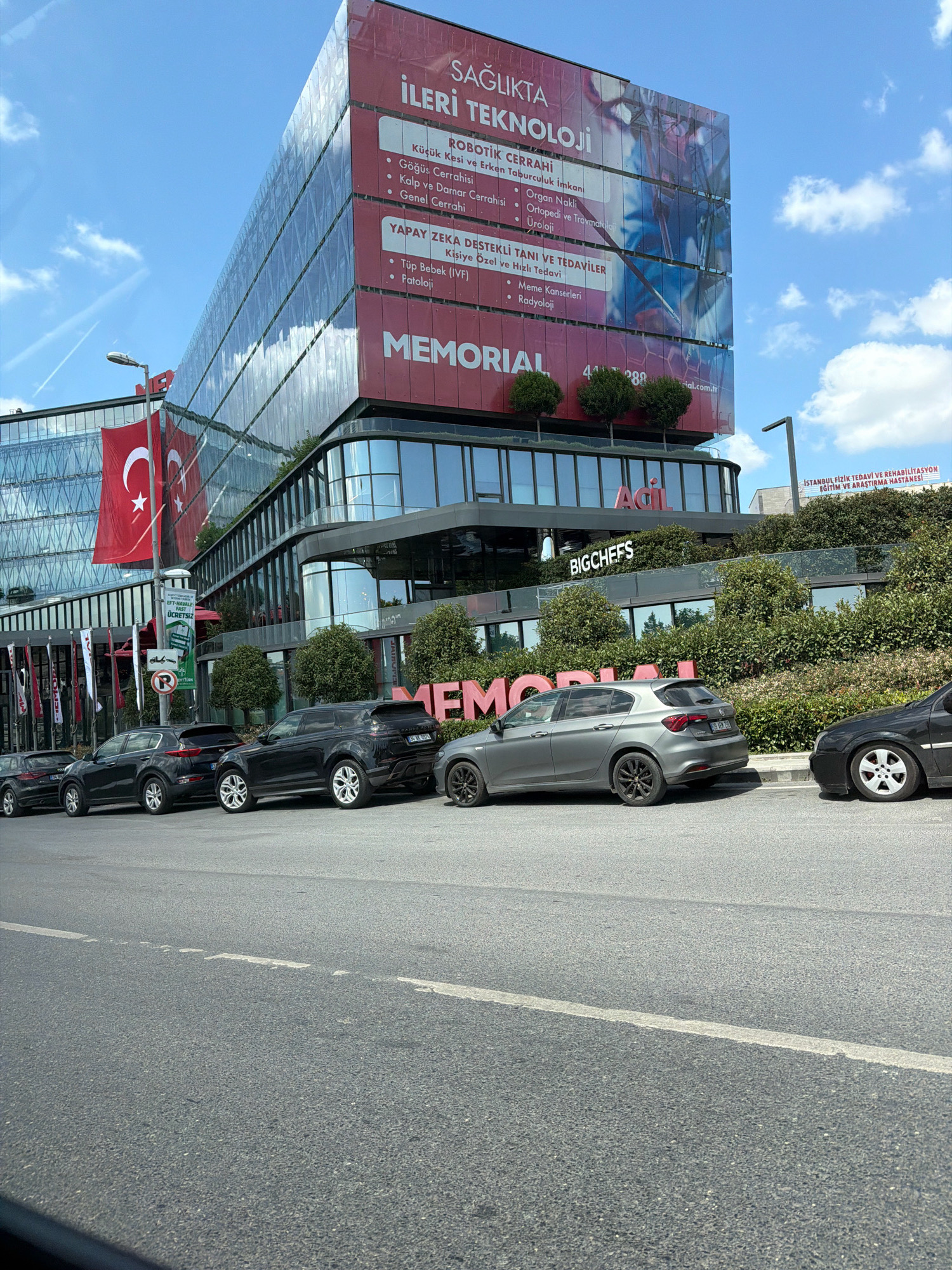 The image shows a modern building with a large red banner displaying "SAĞLIKTA İLERİ TEKNOLOJİ" (Advanced Technology in Healthcare) and "MEMORIAL" in large pink letters. The building appears to be a hospital or medical facility. Several cars are parked on the street in front of the building. A Turkish flag is visible on a flagpole to the left of the building. The scene is set in a city environment with a bright blue sky and scattered clouds. The specific location is Bakırköy, Turkey, as indicated by the context of the image's origin.