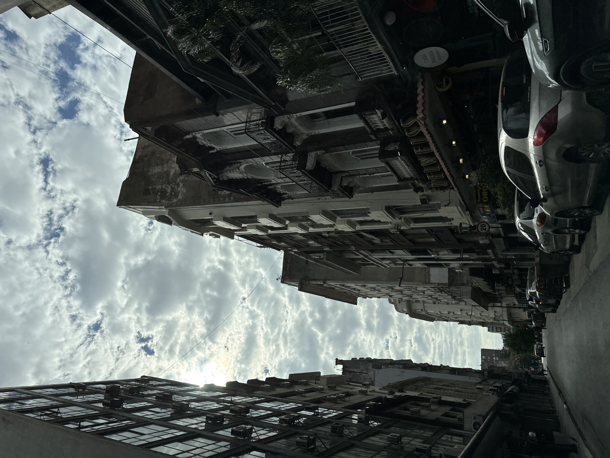 The image, when viewed upright, depicts a narrow urban street looking upwards, creating a "street canyon" effect between tall, multi-story buildings under a cloudy sky. The scene is set during the daytime, with diffuse light indicating either an overcast sky or the sun low in the sky, peeking through the clouds on the left.
The sky is filled with a mix of large, fluffy cumulus clouds and patches of clearer blue sky. Buildings on both sides of the street exhibit varied architectural styles, suggesting different construction periods. Some buildings feature ornate balconies and decorative elements, while others have more modern, grid-patterned windows. Numerous air conditioning units are visible attached to the facades of many buildings. External fire escapes can be seen on a building towards the left. Some dark green foliage, possibly palm trees, protrudes from behind the structures on the right.
The paved street below is lined with several parked cars, predominantly on the right side. A silver hatchback is prominently parked in the mid-ground. No people or overt activity are visible, lending a quiet atmosphere to the urban scene.
Visible text includes a circular sign attached to a building on the right, which reads "CASA ARRENDA" and "15200005". Below it, another sign partially reads "COMPRO ORO" (Spanish for "I buy gold"). A red and white circular street sign, likely indicating a speed limit, shows the number "20". The overall impression is an established urban environment, consistent with the provided context of Buenos Aires, Argentina.