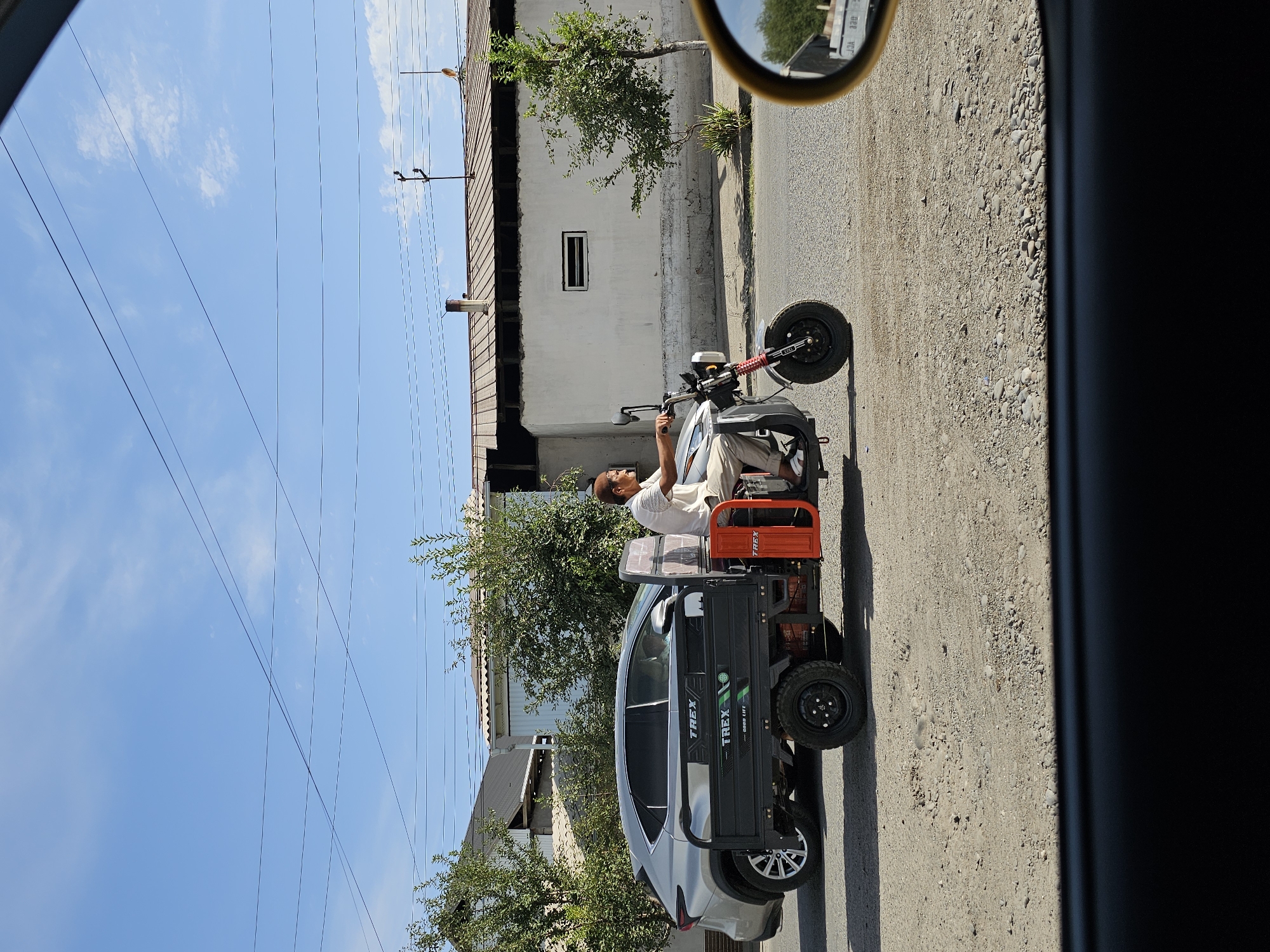 The image shows a man riding a three-wheeled vehicle with a cargo bed. He is wearing a white shirt and pants and a skullcap. The vehicle has a black cargo bed with the brand name "TREX" in white letters. The vehicle is orange and red in some parts. The man is riding on a street with a silver car beside him. The background shows buildings, trees, and a blue sky with some clouds. The street is paved with asphalt and gravel. The image appears to be taken from inside a car, with a black frame at the bottom and a yellow side mirror on the right. The location is identified as Özgön, Kyrgyzstan.