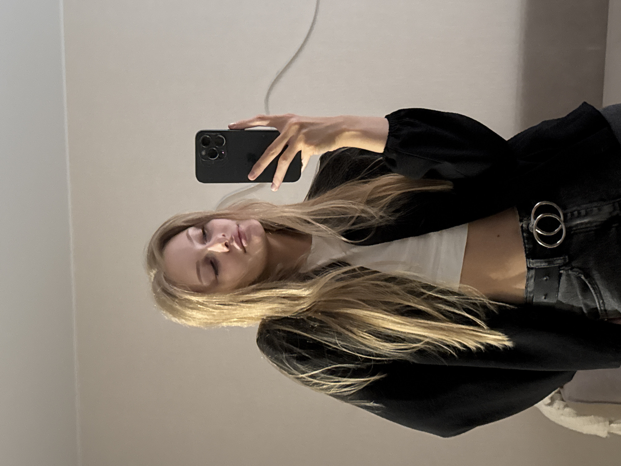 A young woman with blonde hair is taking a mirror selfie indoors. She is wearing a white crop top, a black jacket, and dark jeans with a black belt. The background is a plain wall. The lighting suggests an indoor setting, possibly a bedroom or living area, with a light source creating subtle shadows. There are no distinct indicators of the city of Perm, Russia, within the image itself.