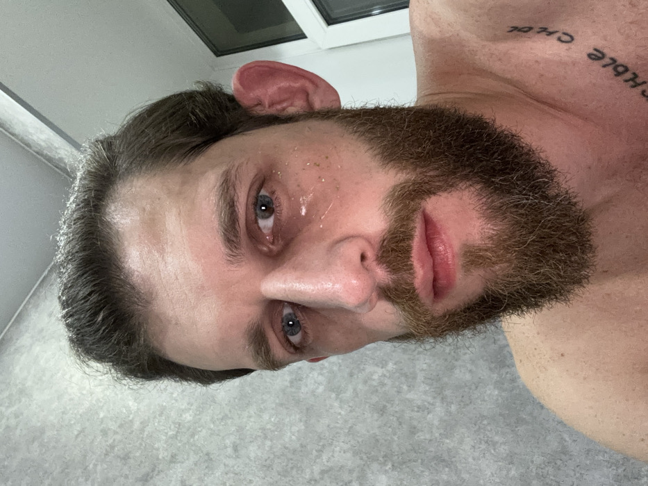 A close-up, tilted headshot of a light-skinned man. He has a thick, brown beard and longish brown hair. His eyes are blue. His face appears slightly wet or shiny. Above his shoulder, a tattoo with text is visible. A window and a textured grey wall are in the background. The image is taken from a low angle, looking up at the man.