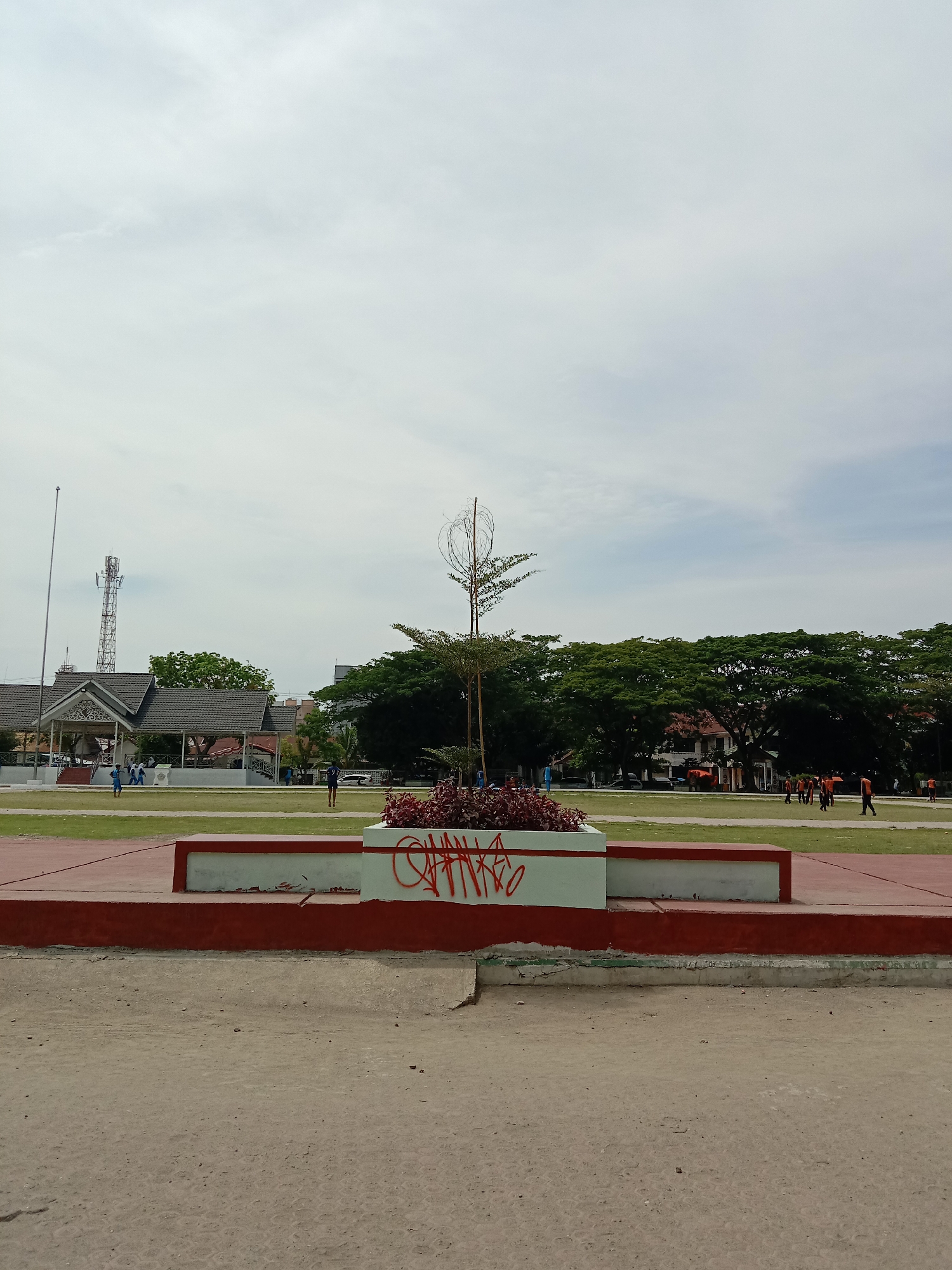 The image shows a public square in Lhokseumawe, Indonesia. In the foreground is a concrete planter box with reddish-purple plants. The planter is vandalized with graffiti. Beyond the planter is a large grassy area where several people are walking and jogging. Some people are wearing blue uniforms, while others are in orange athletic wear. In the background, there's a building with a traditional Indonesian-style roof, a cellphone tower, and a line of large trees. The sky is overcast with light gray clouds. The ground in the immediate foreground is cracked and dirty concrete.