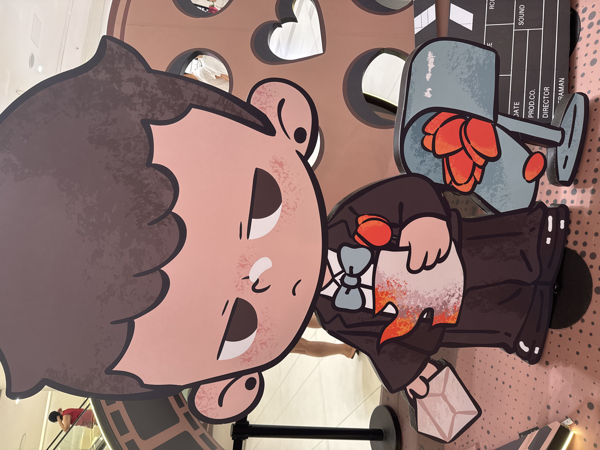 This media file captures a close-up of a large, cartoon-style cutout of a character, likely a boy with brown hair, a light pink face, and large white eyes with black pupils. The character is dressed in a dark suit with a blue bow tie and holds a light-colored object in its right hand and a light brown envelope in its left. The backdrop features cutouts of various shapes, including circles and hearts, behind which are glimpses of reflective surfaces showing people and brightly lit interiors. To the right of the character, a clapboard with visible text is partially obscured. The overall impression is that of a promotional display in a public space, possibly a mall or entertainment venue. The image is oriented vertically, suggesting it might be a portrait orientation photograph. There are no clear indicators of time of day or weather.
Visible text on the clapboard includes: "E", "RC", "SOUND", "DATE", "PROD.CO.", "DIRECTOR", and "-RAMAN".
The scene is likely indoors, given the bright lighting and the presence of what appears to be an escalator and shoppers in the background reflections. The cartoon character's expression is somewhat neutral, with its mouth open slightly and a furrowed brow. The overall aesthetic suggests a fun or creative event. The location context of "Hà Nội, Vietnam" is provided separately, but there are no explicit visual cues within the image to confirm this.