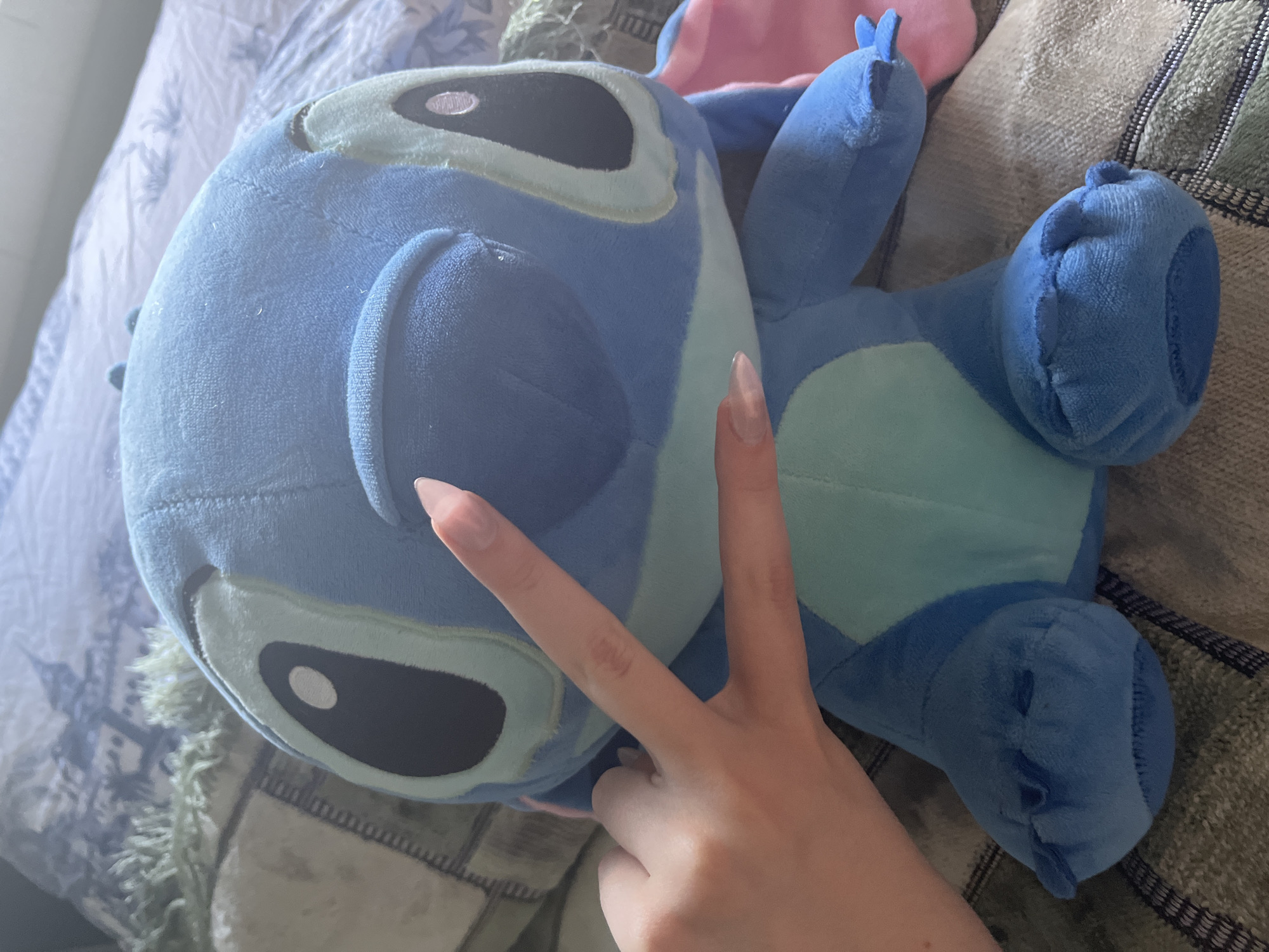 A close-up shot shows a plush blue Stitch toy from Disney's Lilo & Stitch. A person's hand, with long, manicured nails, is positioned in front of the toy, making a peace sign. The Stitch toy is seated on a textured surface, possibly a bed, with a patterned duvet in the background. The background also includes a fuzzy green throw blanket. The scene is indoors, with soft lighting suggesting it's daytime. There is no indication of the city of Ashkadar, Russia.
