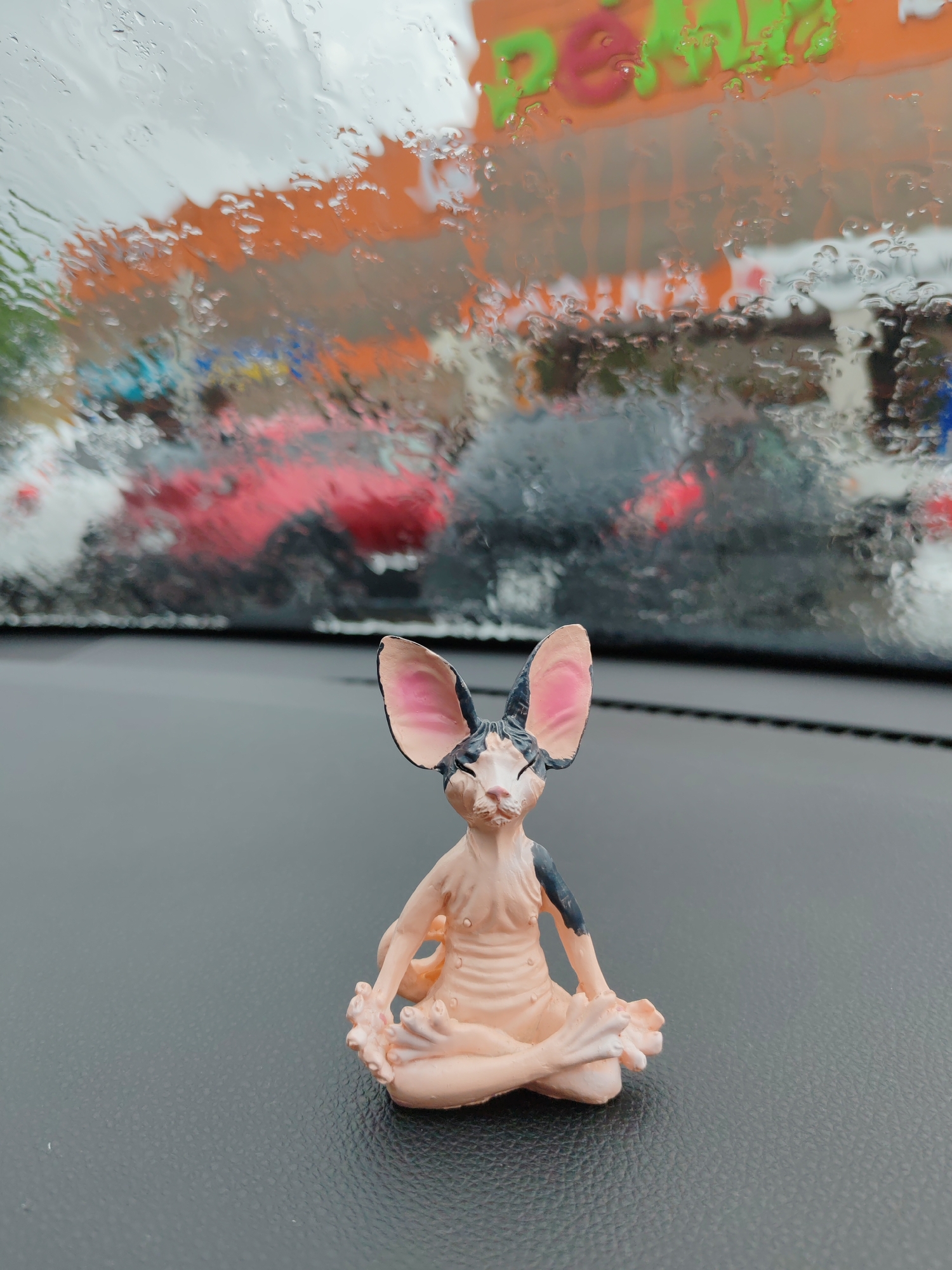 This media file shows a close-up, eye-level shot of a small, figurine of a Sphynx cat meditating. The figurine is light pink with dark grey markings on its ears and shoulders, and it is positioned on a dark grey dashboard in the foreground. The cat is depicted in a cross-legged, seated meditation pose with its eyes closed and hands resting on its knees.
In the background, through a car windshield covered in raindrops, a blurry scene is visible. The raindrops distort the view, creating an abstract impression of what appears to be a street or parking lot. There are hints of colorful buildings with orange and green signage, possibly indicating a commercial area. Red and dark vehicles are also discernible, suggesting traffic or parked cars. The lighting and the raindrops on the windshield imply it is daytime and raining.
The overall mood is serene, due to the meditating figurine, juxtaposed with the slightly chaotic and blurry outdoor environment. The image seems to capture a moment of calm within a potentially busy or mundane setting. There is visible text on the orange signage in the background, which appears to read "PEONY" in stylized green and pink letters. The location context is Komsomolsk-on-Amur, Russia.
