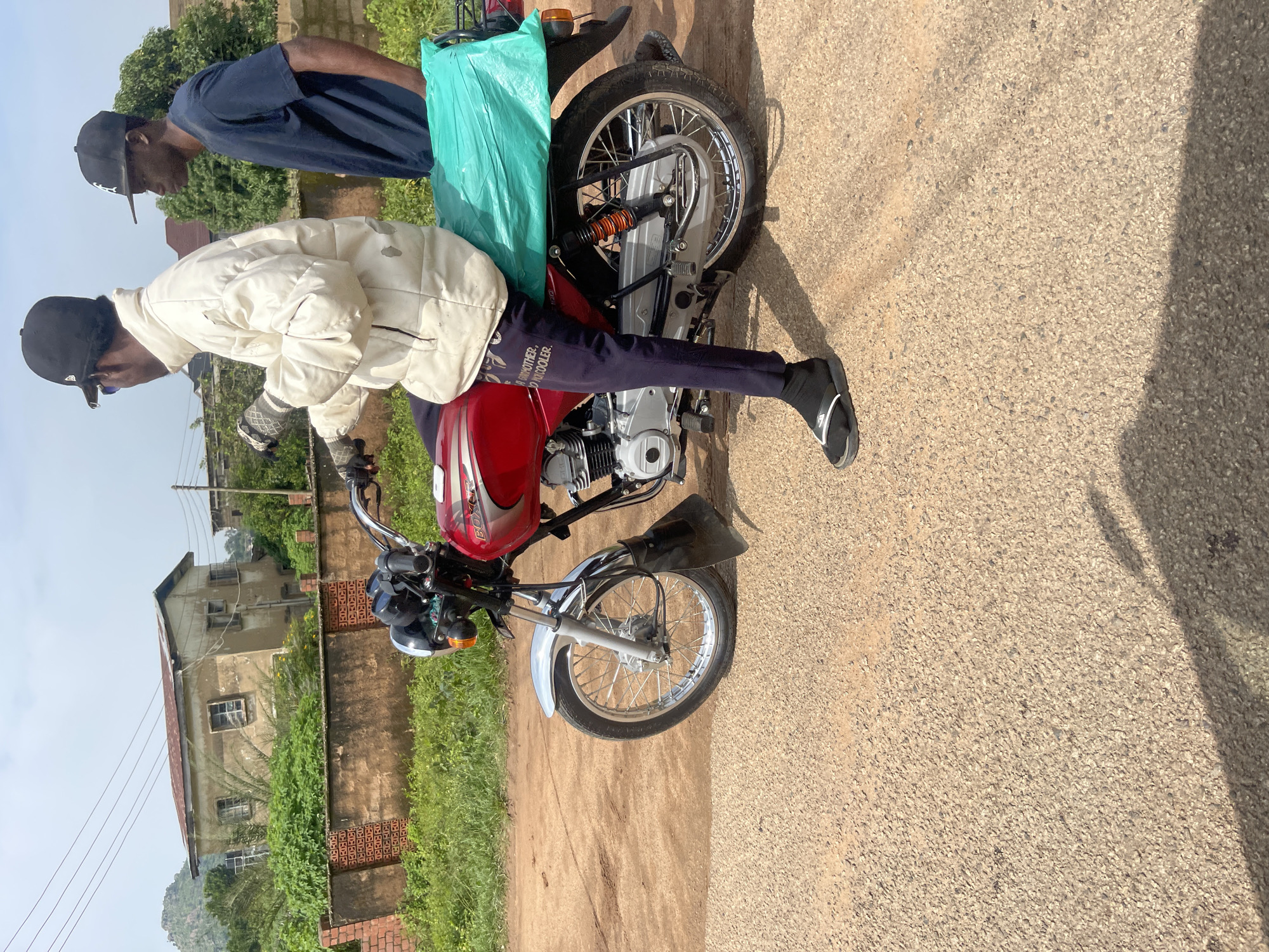 Two individuals, both wearing baseball caps, stand near a red and silver motorcycle parked on a dirt road. The person in the foreground, wearing a white padded jacket and dark pants, is seated on the motorcycle. They have on black flip-flops and gloves. A bright teal bag is strapped to the back of the motorcycle. The second individual, dressed in a navy blue t-shirt, stands behind the motorcycle, bending slightly as if speaking to the person on the bike.
The scene appears to be outdoors, with a building and lush green vegetation visible in the background. The sky is bright, suggesting daytime. The overall impression is a casual interaction or preparation for travel. The image is oriented vertically, with the subjects and motorcycle running from bottom to top.
There is some text visible on the dark pants of the person on the motorcycle, which appears to read "BROTHERHOOD" and "ROCKER". The location is identified as Gwong, Nigeria.