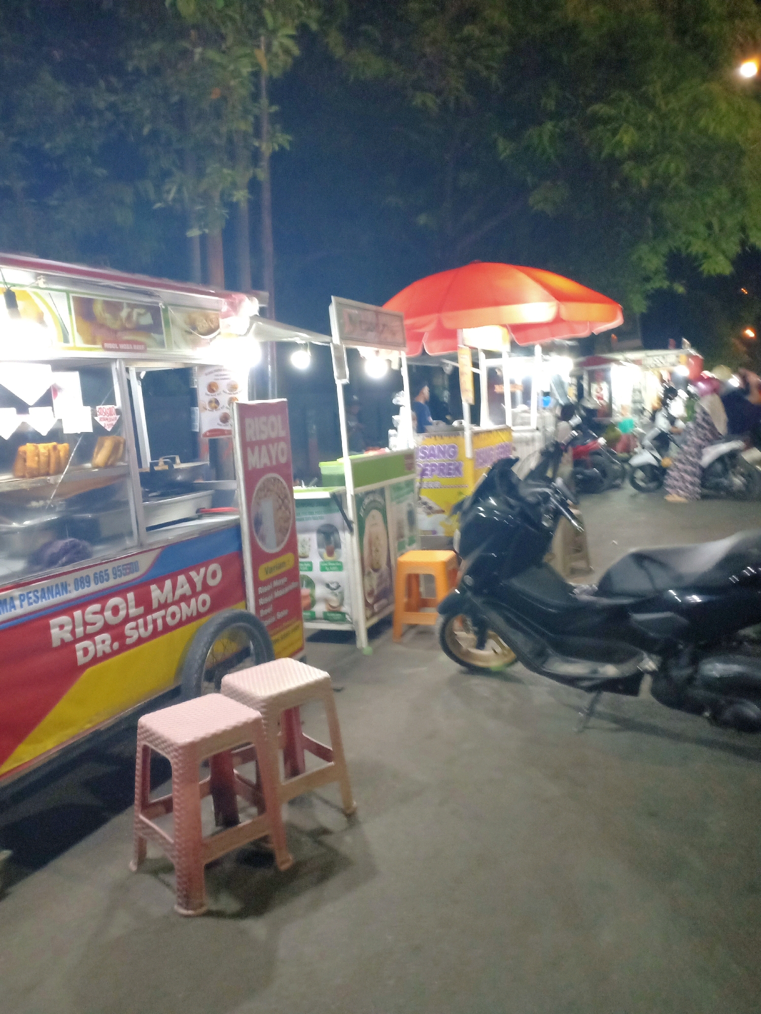 This image depicts a nighttime scene at a street food stall, likely in the city of Tuban, Indonesia, as indicated by the text on the stall itself. The primary focus is a food cart with a prominent banner that reads "RISOL MAYO DR. SUTOMO," suggesting it sells risol mayo. The cart is brightly lit, showcasing food items displayed behind glass. Adjacent to this cart is another stall with an orange umbrella. Several motorcycles are parked in the background, and some individuals are visible, including a person in a patterned hijab and another in a blue shirt. The ground is paved, and trees with lush foliage can be seen in the background, illuminated by the artificial lights of the stalls. Two small, light-colored stools are placed in the foreground. The overall atmosphere is that of a lively evening marketplace.