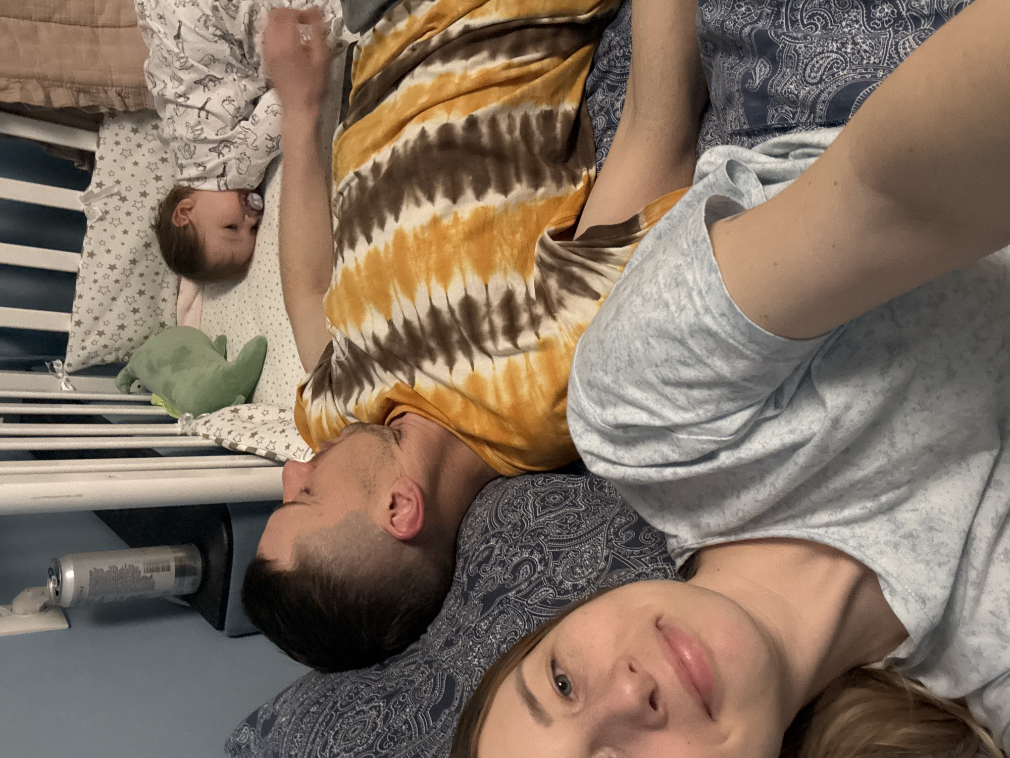 A family of three is lying together on a bed in what appears to be a bedroom. A woman is in the foreground, taking a selfie. A man lies behind her, next to a baby who is asleep in a crib.
The woman has light skin, blond hair, and is wearing a light blue shirt. She appears to be lying on a blue paisley patterned pillow. The man has short, dark hair and is wearing a tie-dye shirt. The baby is wearing a white onesie with animal prints and has a pacifier in its mouth. A stuffed green dinosaur is next to the baby.
The crib is white with horizontal bars. A can and an electrical outlet are visible on the wall behind the crib. The location is most likely a domestic setting, and not Moscow, Russia.