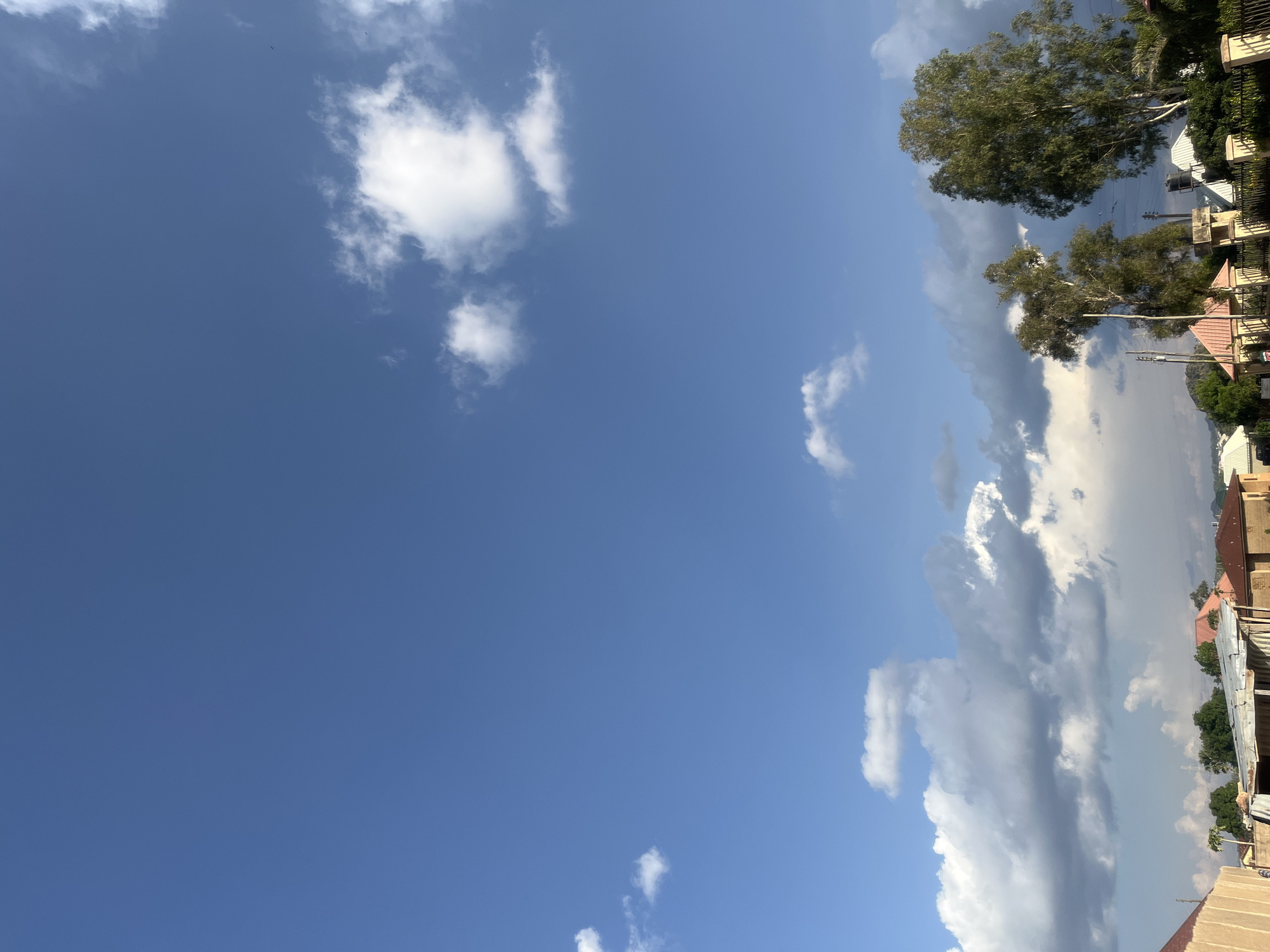 The image captures a residential landscape in Barakin Akawo, Nigeria, under a bright, partly cloudy sky. The upper two-thirds of the frame consists of a clear blue sky, interspersed with scattered white cumulus clouds. Small, dark specks, possibly birds, are visible in the upper right quadrant. Below the clear sky, a band of larger, denser greyish-white cumulus clouds stretches across the middle of the frame, indicating varying cloud formations.
The lower portion of the image reveals a collection of one-story residential buildings. These structures primarily feature reddish-orange or reddish-brown pitched roofs, likely constructed from corrugated metal, and walls rendered in light beige or off-white plaster. One building in the lower-middle left has a silver corrugated metal roof. On the right, a black cylindrical water tank is mounted on a white rooftop. The foreground includes various walls and fences, some with decorative pillars.
Multiple trees with green foliage are present throughout the scene. Two prominent, tall trees with lighter-colored trunks stand on the right side of the frame, dominating that section of the view. The sunlight suggests either a morning or late afternoon setting, casting distinct illumination on the clouds and structures. No individuals or discernible activities are observed.