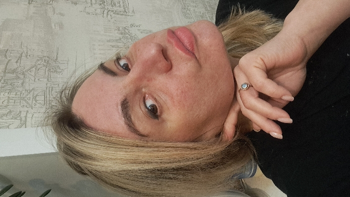A female individual is depicted in a close-up indoor portrait, presumed to be a selfie, located in the city of Bel'kovo, Russia. The subject is an adult female with blonde hair, appearing lightened with darker roots and some loose strands. Her skin tone is light, exhibiting visible pores and localized redness on the nose and cheeks. Her dark brown eyes are directed towards the camera, with a thin line of dark eyeliner on the lower lash line. Her eyebrows are dark and shaped. Her lips are full, colored with light pink lipstick, and positioned in a puckered expression. She wears a black garment, visible on her shoulder and chest. Her left hand is positioned with fingers resting against her chin and jawline; the visible fingernails are manicured with a pale, natural polish. A ring with an oval, light-colored stone, possibly opalescent, is present on her left ring finger. The background consists of a wall covered with wallpaper featuring a monochromatic, illustrated urban cityscape design, including various architectural structures and faint outlines of vehicles. A blurred green object, consistent with foliage, is visible in the upper left quadrant of the image. The scene is illuminated by direct, likely artificial, light.