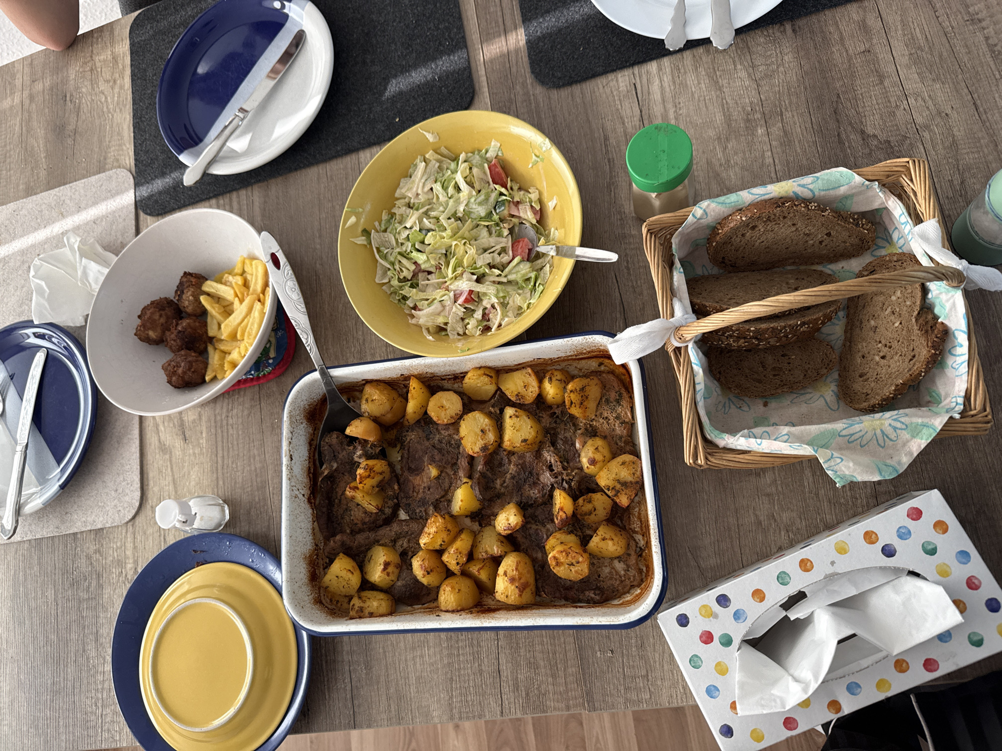 The image depicts a table laden with food, possibly in Bruchköbel, Germany, though no specific landmarks are visible. A rectangular baking dish filled with roasted meat and potatoes sits centrally. A yellow bowl contains a salad, and another white bowl holds meatballs and French fries. Bread is presented in a wicker basket lined with a floral cloth. Blue and white plates, cutlery, and a tissue box with colorful polka dots are also visible on the wooden table. No people are discernible in the image.
