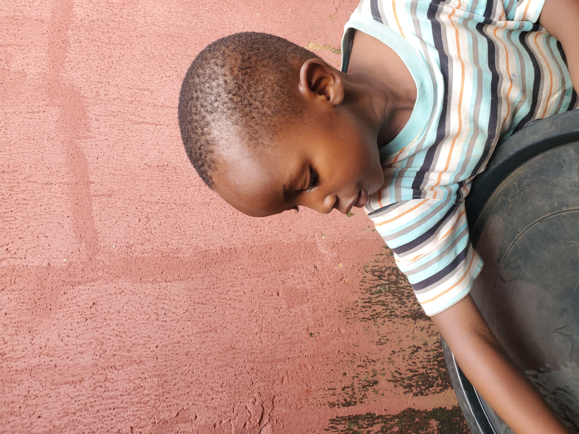 The image shows a close-up, high-angle view of a young Black child lying on a textured, reddish-brown surface, possibly a wall or ground with a rough plaster finish. The child is wearing a striped t-shirt with horizontal bands of white, light blue, black, and orange. The child's head is turned downwards, with eyes closed or looking down, suggesting a state of rest, contemplation, or sleep. The child's right arm is visible, resting on a dark, circular object, which appears to be a bucket or a large container. The overall lighting suggests it might be daytime. The textured surface on the left side of the image is consistent with outdoor environments in locations like Jos, Nigeria, where such finishes are common. The scene conveys a sense of quietness and simplicity. There is no visible text in the image.