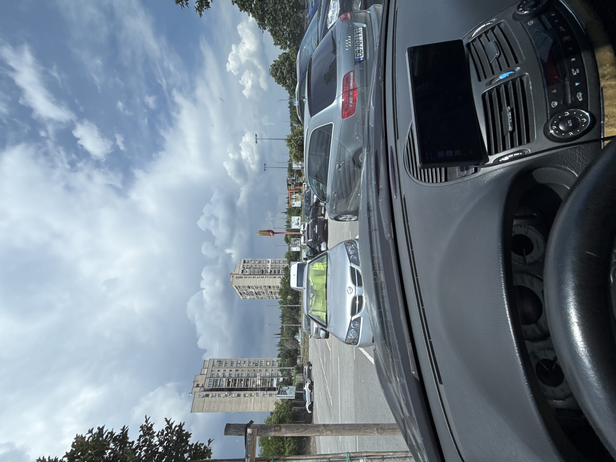 The image depicts an outdoor urban scene in the city of Stara Zagora, Bulgaria, captured from the interior of a vehicle.
In the immediate foreground, the dashboard of the viewer's car is visible, featuring dark grey textured plastic. A portion of a black steering wheel is present at the bottom left. Partially obscured instrument gauges, including a speedometer marked up to 260 km/h, are visible. The central console features air vents, climate control buttons (e.g., "AUTO," "AC REST"), and rotary knobs. A black rectangular electronic device, likely a smartphone or tablet in a mount, is positioned atop the central air vents, displaying a dark screen with faint icons and a digital time of "12:43." A connecting cable extends from the device.
Beyond the windshield, a paved asphalt parking lot unfolds under a partly cloudy sky, showing patches of blue and significant grey cloud formations. Directly ahead, a silver Nissan sedan is parked, its windshield covered by a bright yellow-green sunshade. To its right, a grey Audi A6 C6 Avant (station wagon) is parked, displaying a Bulgarian license plate "CB 7071 XT." Further back in the parking lot, additional vehicles are visible, including a white sports car and a dark SUV on the left, and a white commercial van behind the Nissan.
In the mid-ground and background, two tall, multi-story concrete residential buildings, characteristic of block apartment architecture, dominate the skyline. Between these buildings, the distinctive red and yellow "M" logo of a McDonald's restaurant is visible. Green trees and dense vegetation line parts of the parking lot perimeter and are present in the far background. Several streetlights and utility poles are also scattered throughout the scene. No individuals are discernible within the frame.