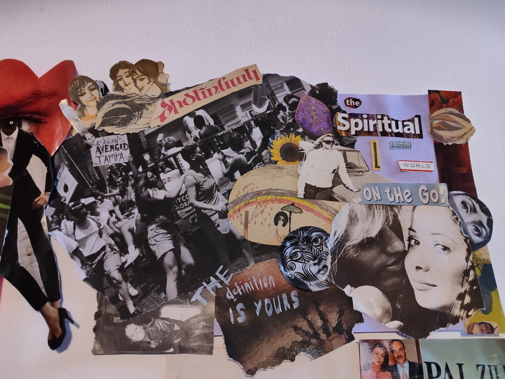 This is a collage of various cut-out images assembled on a white wall. The collage includes several black and white photographs of people, including scenes that appear to be from a march or gathering, and portraits of individuals. There are also text snippets incorporated, such as "LESBIAN'S AVENGED TAMPA," "Spiritual EASTERN WORLD," "ON the Go!" and "THE definition IS YOURS." Additional artistic elements include colored cut-outs, like a red heart, a sunflower, and a face illustration. A photograph of a couple and the partial word "DAI Z" are at the bottom. There is no evidence to suggest that this collage is located in the city of Gyumri, Armenia.