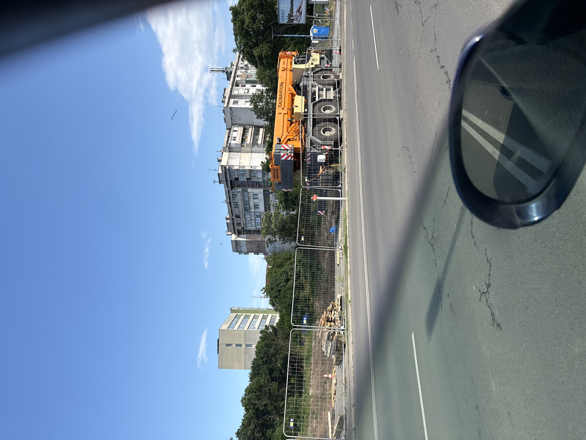 The image captures a construction site in Burgas, Bulgaria, viewed from a vehicle. In the foreground, a portion of a car's side mirror is visible, reflecting the road ahead which has white lane markings and some cracks. A bright orange crane, identified by Cyrillic text and "GROVE" branding, is the central focus of the construction site. The crane is positioned behind a metal safety fence, along with stacks of lumber, construction materials, and a portable blue toilet. Beyond the fence, a tall, grey, multi-story building stands, and further in the background are more apartment buildings and lush green trees. The sky above is a clear blue with a few scattered clouds, and a bird is visible in flight. The overall scene suggests ongoing urban development or renovation.