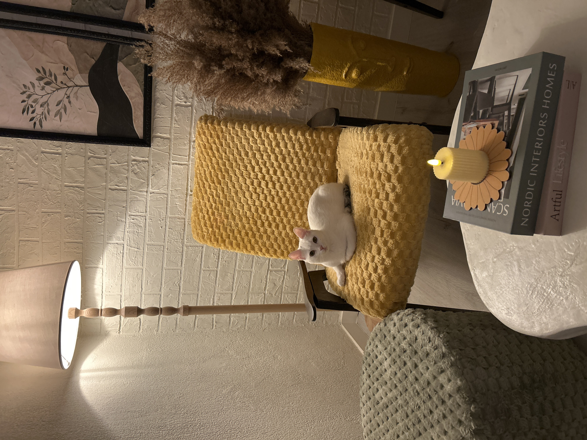 A white cat is lying on a yellow fuzzy armchair in a room. The room has white brick walls, a tall floor lamp, a green pouf, a coffee table with books and a lit candle, and a yellow vase with dried plants. Two framed pictures of minimalist art hang on the wall.