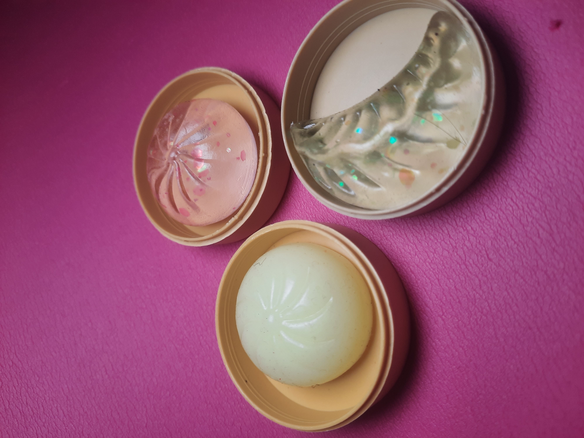 Three open cosmetic containers are arranged on a textured magenta surface. The container on the left holds a pale green, spherical object with a pleated pattern on its surface. The container in the upper right contains a translucent pink object with a similar pleated pattern, studded with small, glitter-like particles. The container in the lower right holds a clear, gel-like substance with green and iridescent glitter. This image does not depict any identifiable location in North Jakarta, Indonesia.