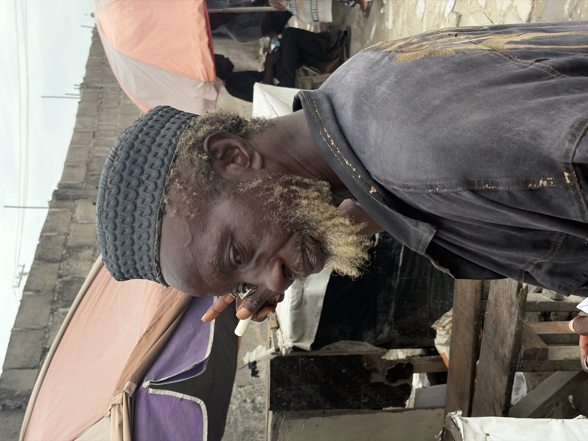 A middle-aged African man in Igboefon, Nigeria, is smoking a cigarette. He wears a dark gray textured kufi and a matching jacket. His beard is sparse and has gray patches. He holds the cigarette in his right hand, close to his mouth. The background is cluttered with elements of a marketplace, including tents with purple and tan fabric and exposed wooden beams, suggesting semi-permanent stalls. Other people are discernible in the background, some sitting. The setting appears to be outdoors, with overcast sky. The man's gaze is downward and to the left. The environment seems to be a street with concrete structures visible in the distance.