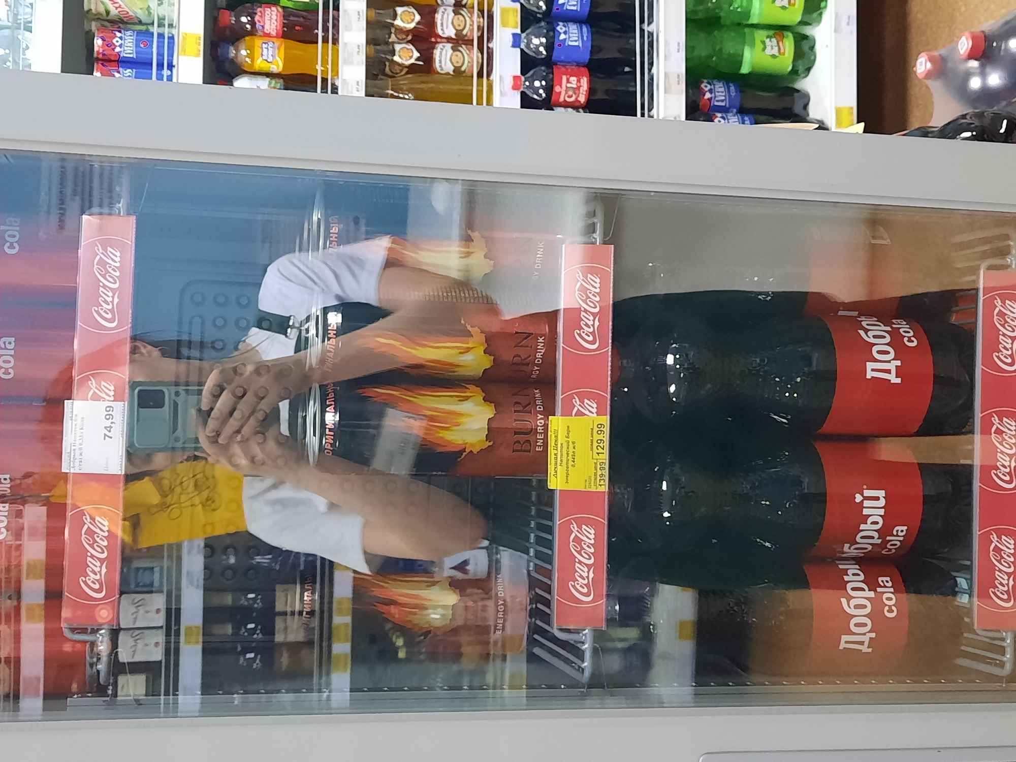 The image shows a person reflected in a refrigerator door in what appears to be a grocery store. The shelves of the refrigerator are stocked with various beverages, including Coca-Cola, "Dobry" cola (a Russian-market Coca-Cola variant), Burn energy drink, and bottles of juice from "Everyviss". The shelves of the refrigerator are in a commercial setting, and the brands of the beverages for sale are recognizable. The person reflected in the door appears to be taking a picture with a smartphone.
Additional details provided by OCR in the image indicates a pricing label for Coca-Cola at 74.99 and for Burn energy drink at 139.99, with a sale price of 129.99. Text on a package of soft drink product says "Original". Based on the Cyrillic lettering on the "Dobry" cola bottles, the image was most likely taken in Russia or a region where Russian is commonly spoken.
The location is in Krasnogorskoye, Russia.