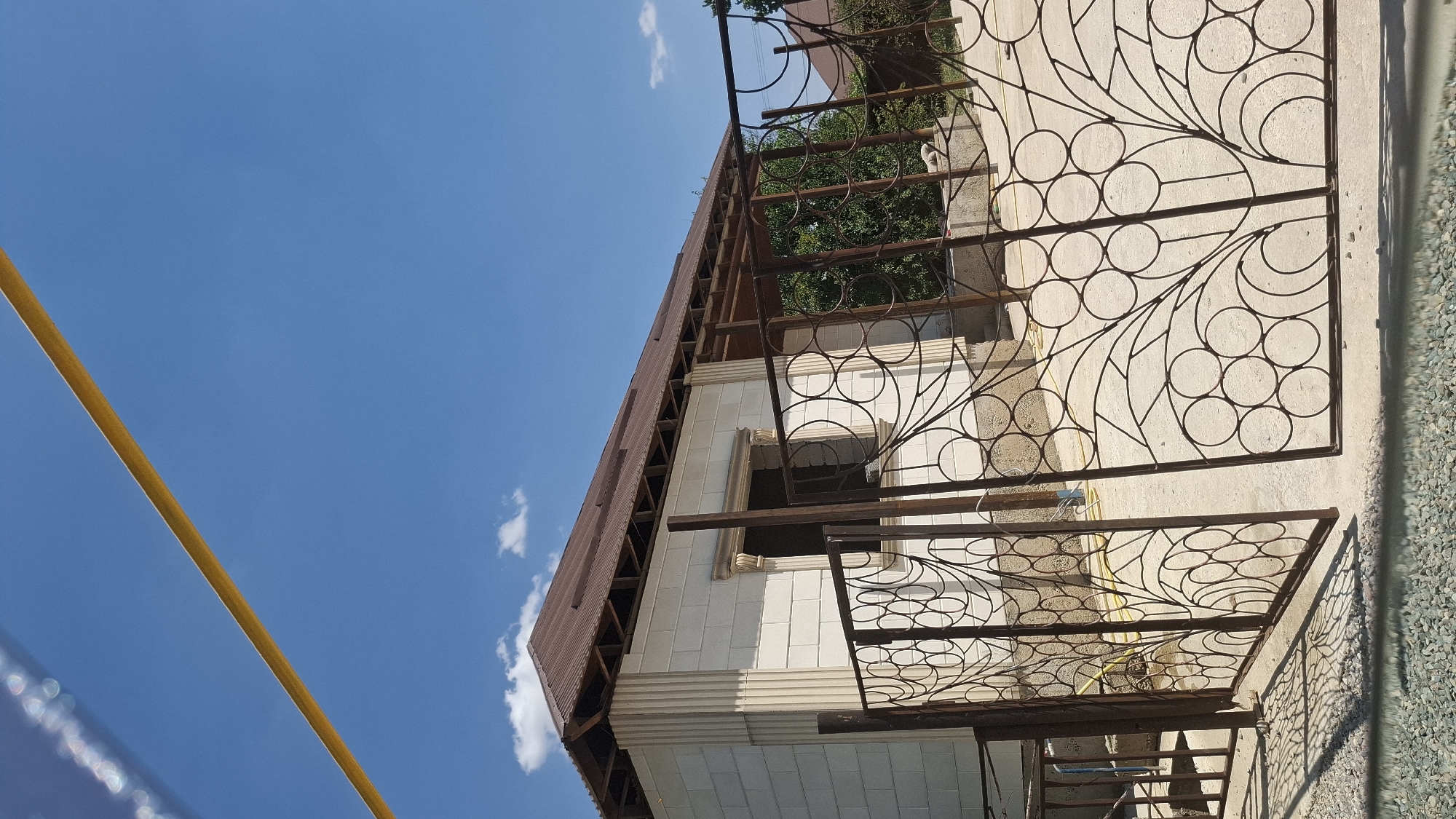 The image shows a house under construction, with a brown metal roof and white brick walls. The house features decorative columns on the exterior and an open porch area supported by wooden beams. In the foreground, there is an ornate wrought-iron gate with a circular pattern. The ground is a concrete surface, with some gravel visible in the lower part of the frame. A yellow pipe or hose runs across the concrete. The sky is blue with scattered white clouds. Trees are visible in the background, suggesting a suburban or rural setting. The image appears to be taken in daylight. The location is identified as Shubarssu, Kazakhstan.