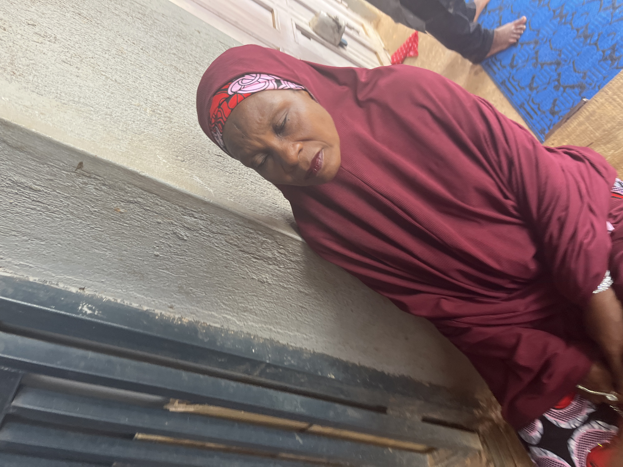 A woman wearing a maroon hijab and robe sits with her eyes closed, leaning against a textured gray wall. She has on a headscarf with a red and pink floral pattern. To her left is a dark, ribbed metal door or gate. To her right, a blue mat with a black pattern is visible on the ground, and a portion of another person's legs and feet in black pants is seen standing near the mat. The environment appears to be outdoors, possibly in an alleyway or near a building entrance. This scene is located in the city of Jos, Nigeria.
