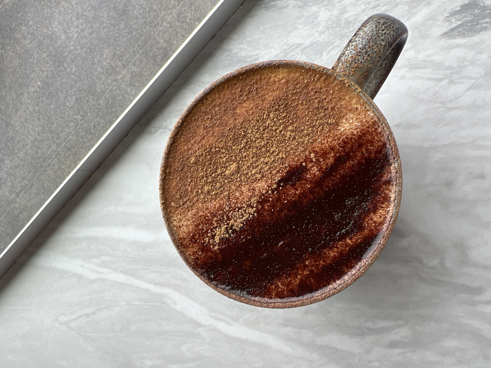 A ceramic mug filled with a hot beverage, likely coffee or a latte, sits on a marble surface. The drink is topped with a swirl of chocolate syrup and a dusting of cinnamon. The mug has a speckled, rustic glaze and a C-shaped handle. In the background, a thin metal strip runs diagonally, separating the marble surface from a grey, textured surface. The scene is lit by natural light, casting subtle shadows. The setting appears to be indoors, possibly a cafe or home kitchen. There is no indication of the city of Donegal, Ireland, within the image.
