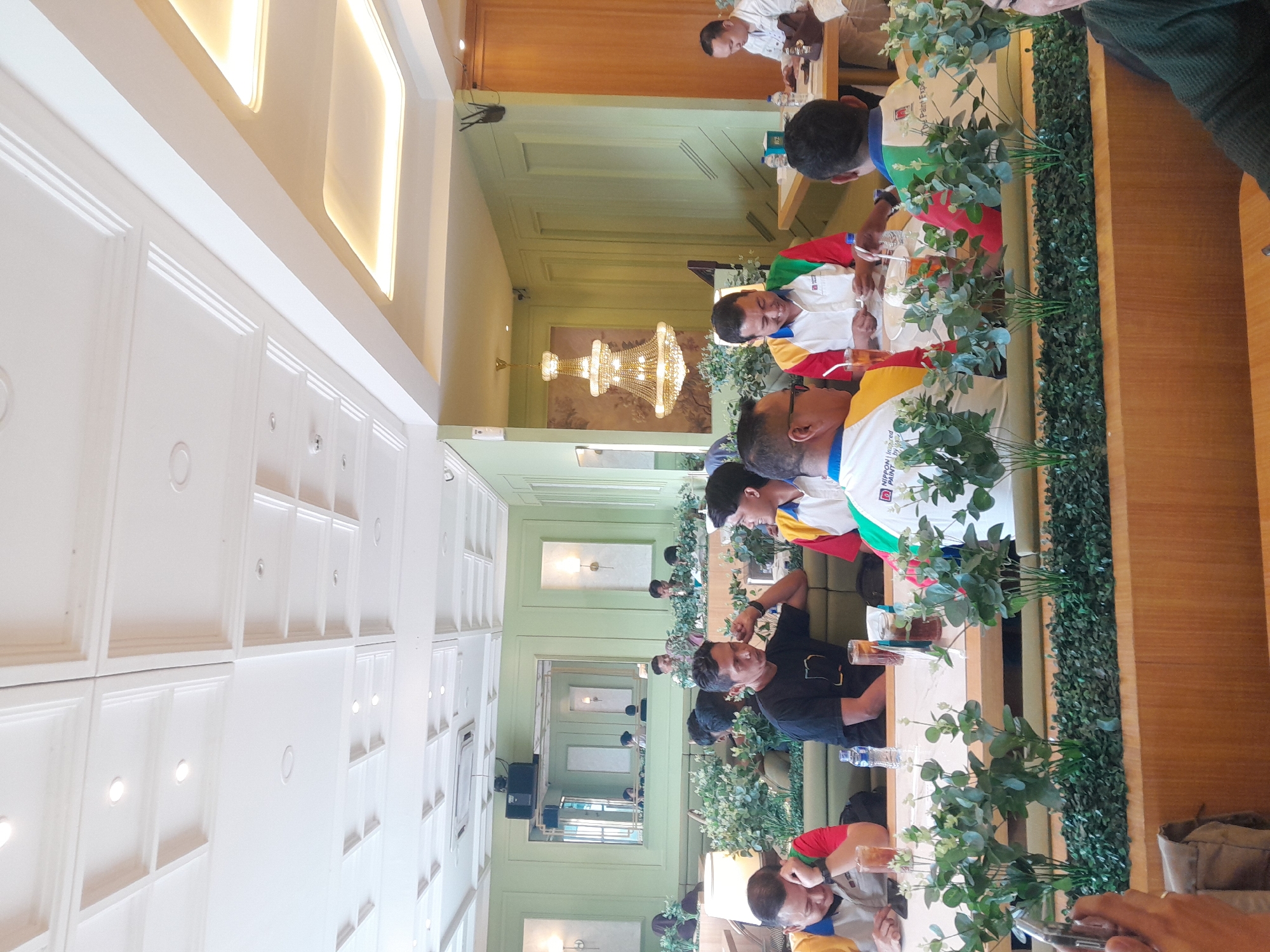 The image depicts an indoor scene, likely a restaurant or cafe, with a group of men seated at a long table. The table is set with drinks, including iced tea in glasses and bottles of water. The men appear to be engaged in conversation or a meeting, with some looking at each other and others looking down at the table.
The environment features a light green wall, a decorative chandelier, and a large mirror reflecting the room. The ceiling has a coffered design with recessed lighting. There are also artificial plants placed along the edge of the table, adding a touch of greenery.
One man on the left is wearing a shirt with red, yellow, and blue accents. Another man in the center wears a black t-shirt. Further down the table, several men are wearing shirts with colorful panels of red, yellow, green, and blue, some with what appear to be company logos. A man on the far right is wearing a light-colored button-down shirt. The overall atmosphere suggests a casual gathering or a business meeting in a well-decorated indoor space. The location is stated to be in the city of Sempusari, Indonesia.