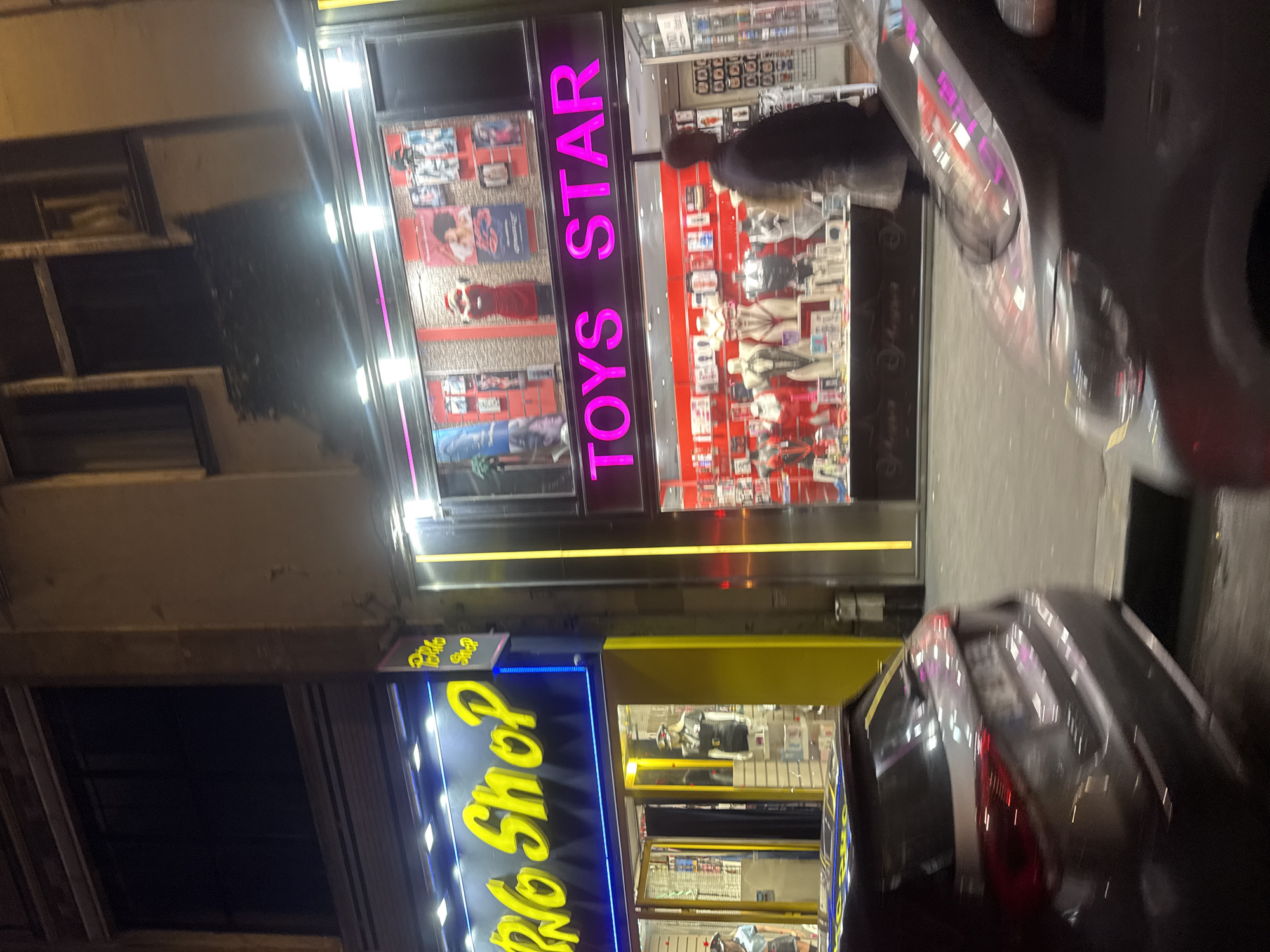 The image shows the exterior of two adult shops in Paris, France, at night. On the left, "Toys Star" is illuminated with pink neon lettering, displaying lingerie and novelty items in its window. Next to it, "Bikini Shop" has a blue and yellow neon sign, showcasing swimwear. A man is seen inside "Toys Star." Cars are parked along the street, partially obscuring the storefronts. The buildings appear to be older with traditional Parisian architecture.
