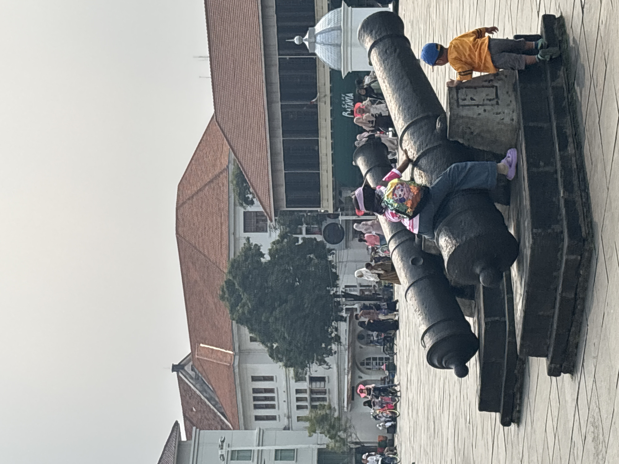 The image shows two cannons on display in a public square in West Jakarta, Indonesia. A young girl with a backpack sits on the larger cannon, facing away from the camera, while a young boy in an orange shirt stands on the base of the cannon, reaching out towards it. In the background, several people are walking around, and there are buildings with red tiled roofs. One building has a sign that reads "CAFE BATAVIA." The ground is paved with square stones. The sky is overcast.
