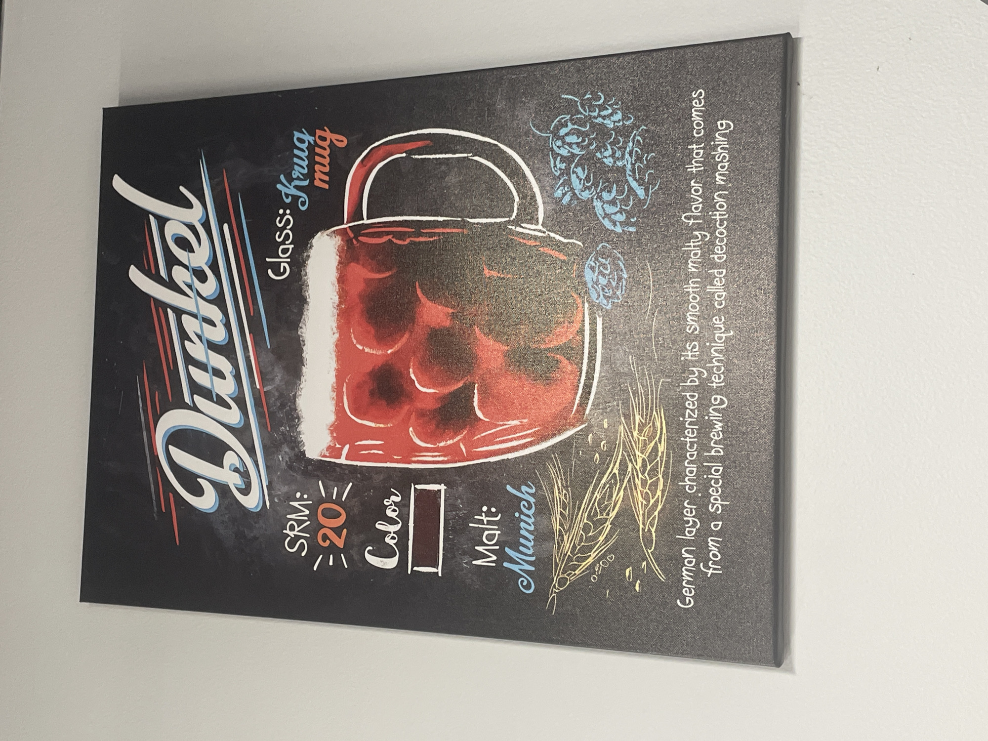A black sign with white, red, and blue lettering advertises "Dunkel" beer. The sign features an illustration of a dark red beer in a mug, along with depictions of wheat stalks and hop cones. Text on the sign provides details about the beer, including its SRM (20), color (a dark brown rectangle), malt type (Munich), and the glass it is served in (Krug mug). A description of the beer's flavor and brewing technique is also present. The sign is mounted on a white wall.