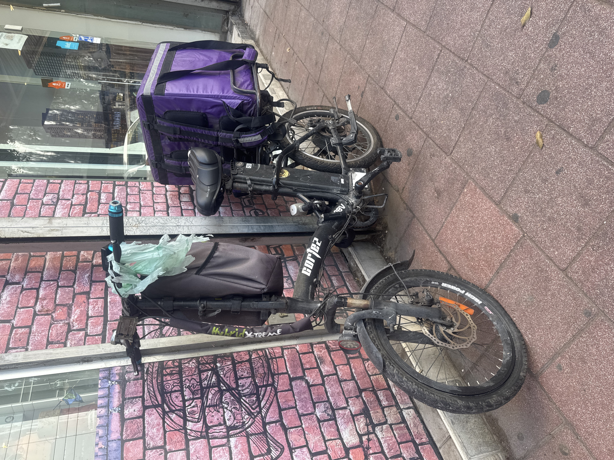A black folding electric bike, with a purple delivery bag strapped to the back, leans against a storefront in Ramat Gan, Israel. The bike has a flat front tire, a gray bag attached to the frame, and plastic bags hanging from the handlebars. The storefront has a brick pattern and graffiti on the lower portion. The sidewalk is paved with reddish-brown brick.