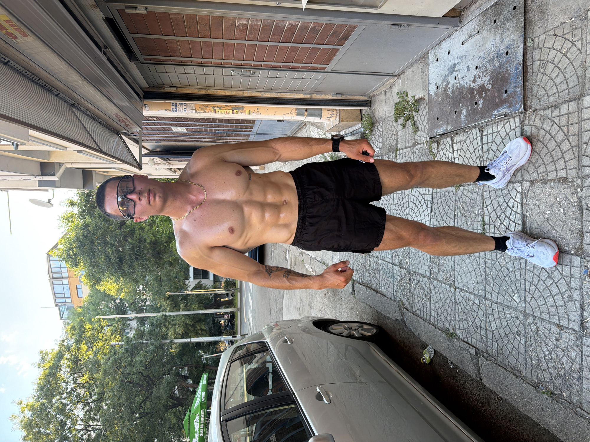 A shirtless, muscular man wearing black athletic shorts, white athletic shoes with red accents, and dark sunglasses stands on a tiled sidewalk in front of a building with metal roll-up doors. He has a gold chain around his neck and a tattoo on his left arm. To his left, a silver car is parked on the street. Trees and other buildings are visible in the background under a clear sky. The man is looking towards the camera. The scene appears to be on a street in the city of Plovdiv, Bulgaria.