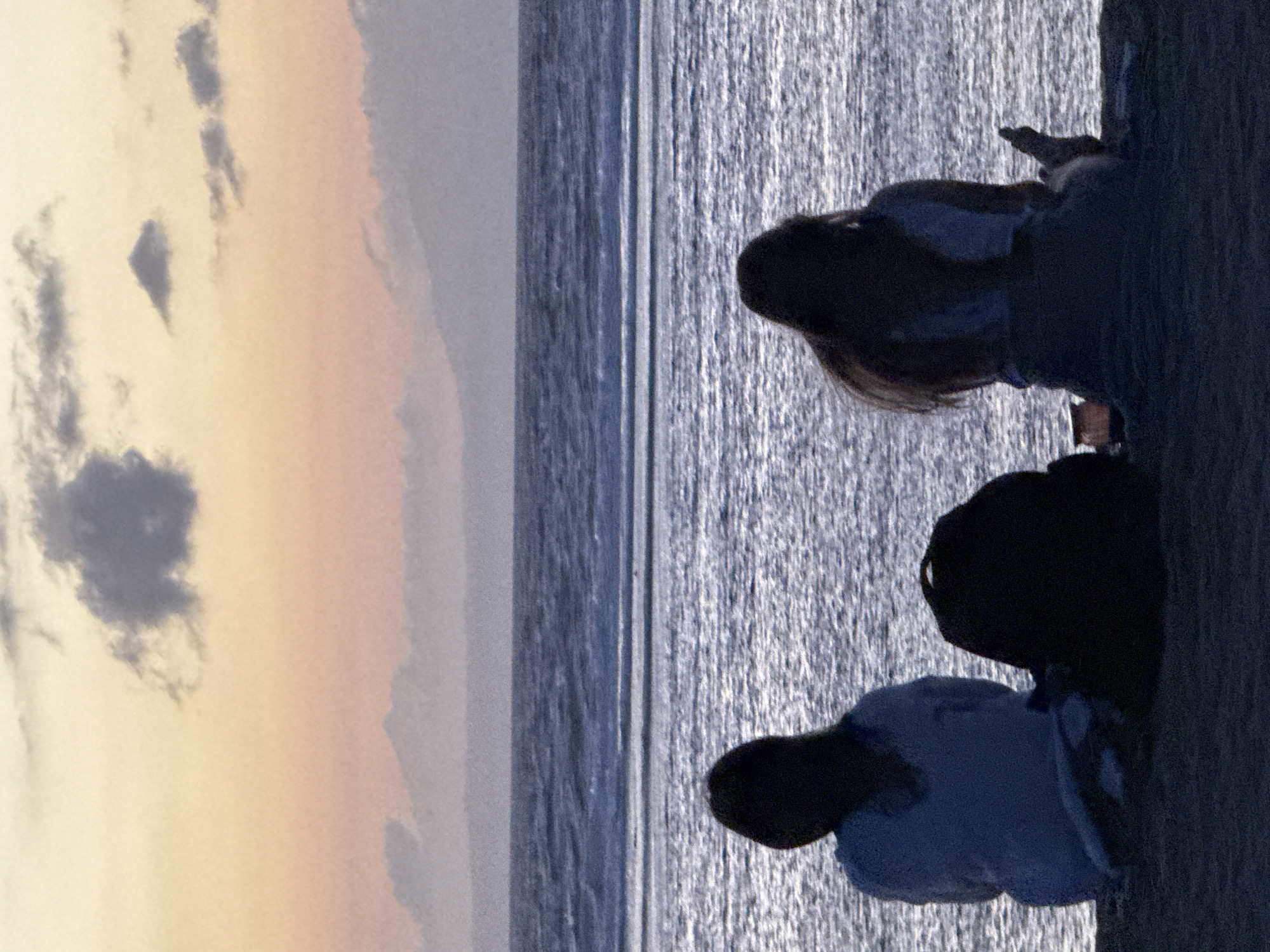 A serene photograph, oriented horizontally, captures three silhouetted individuals observing a tranquil ocean scene, likely during either sunrise or sunset. The sky, in the upper left, displays soft gradients of pale orange and pink, transitioning to a light blue higher up, with scattered, soft-edged clouds catching the warm light.
The middle and right side of the image are dominated by the vast expanse of water, reflecting the ambient light with subtle textures of blue and gray, suggesting a calm sea. On the far right, the three figures, seen from behind and in dark silhouette, are seated or kneeling on a dark, undefined foreground surface, looking out towards the horizon. Their posture suggests they are at rest, contemplating the view.
The scene evokes a peaceful and contemplative mood. The visual elements are consistent with a coastal environment, such as Kuta Selatan, Indonesia. There is no visible text in the image.