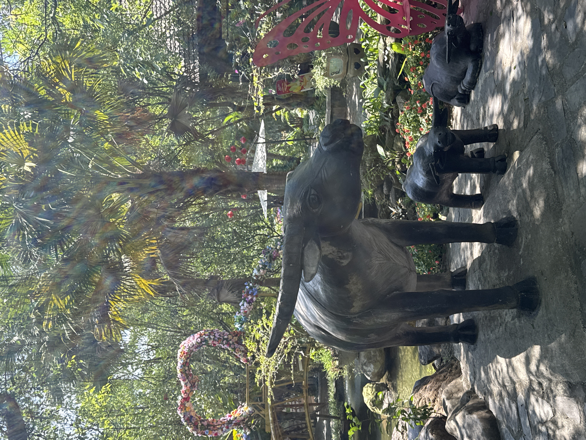 The image captures a scene in what appears to be a garden or park. The primary subject is a weathered, stone statue of an elephant, positioned prominently in the foreground, and slightly behind it are two smaller statues of elephants. The ground is a mix of stone and dirt, with signs of vegetation. In the background, there is a floral heart-shaped decoration and lush foliage, including palm trees and other greenery. The overall lighting suggests a sunny day.
While the specific location is not directly identifiable from the image, the prompt asks for the city of Đà Nẵng, Vietnam. Given the garden setting with specific flora and stone art, it is plausible that this is within a park or botanical garden located in Đà Nẵng, Vietnam, specifically the Marble Mountains or a similar tourist location.