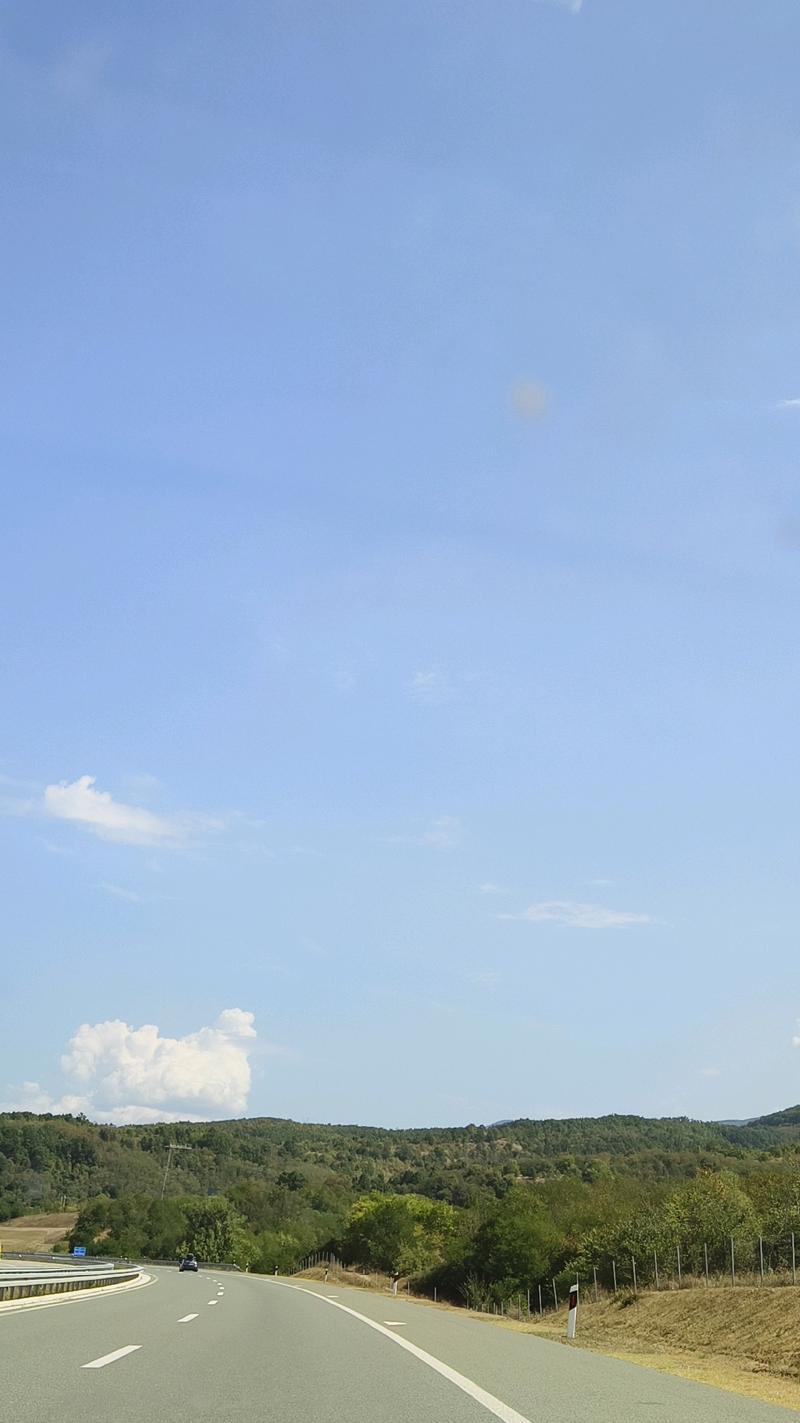 This is a vertical, low-angle, outdoor photograph of a highway curving through a rural, hilly landscape under a clear blue sky with scattered clouds. The highway, paved with asphalt, has white lane markings. On the left side of the highway, there is a concrete barrier. In the distance, a dark-colored car is driving on the highway. The landscape features a dense forest of green trees and shrubs covering rolling hills. On the right side of the highway, there is a dry, grassy embankment with a fence made of thin posts. The scene appears to be located on the outskirts of Vranjska Banja, Serbia. The lighting suggests it is daytime, possibly late morning or early afternoon.