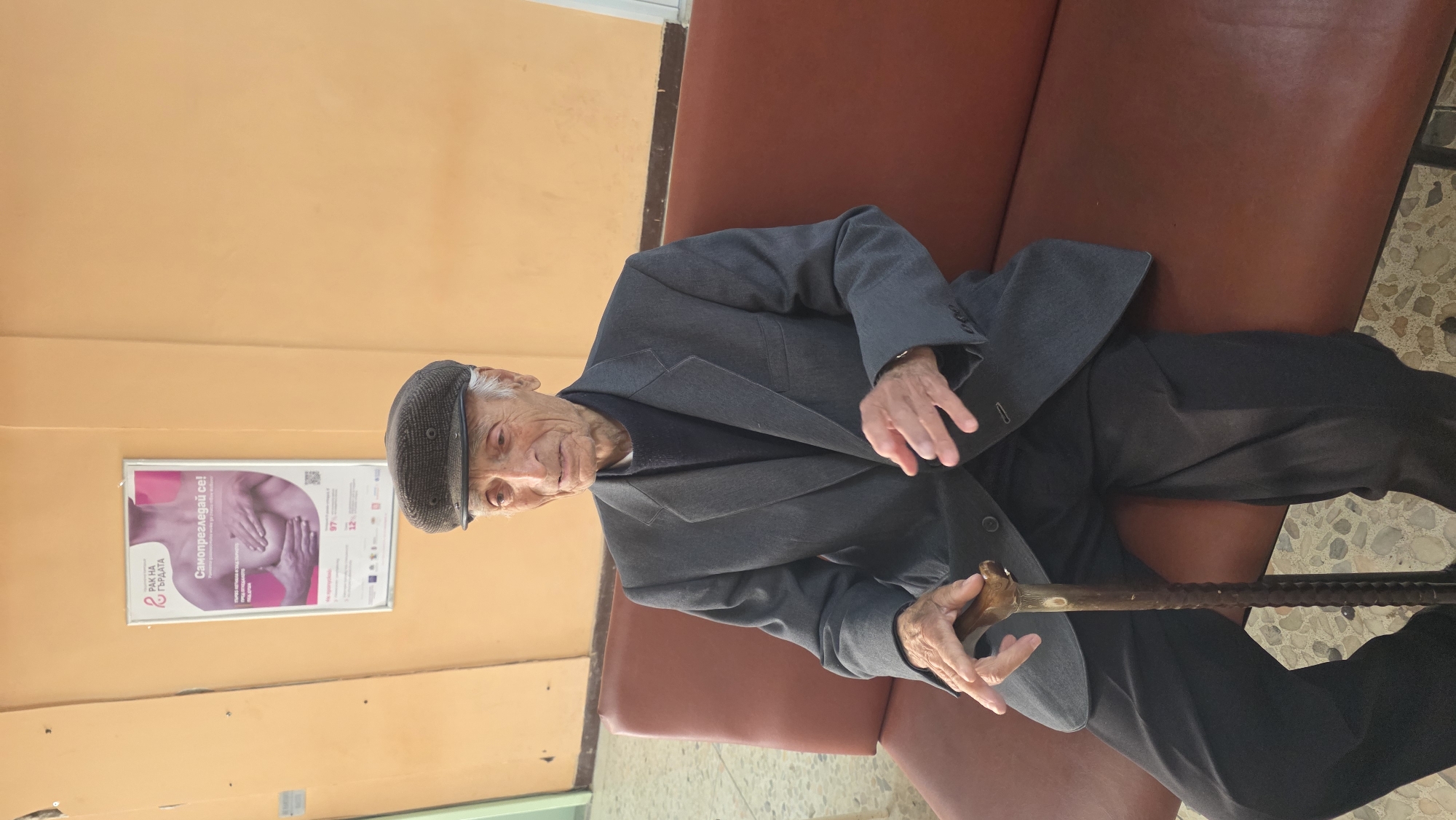 An elderly man sits on a red-brown couch in what appears to be a waiting room in Blagoevgrad, Bulgaria. He is dressed in a dark gray suit and a cap. He holds a cane in his hands. A poster with Bulgarian text and a graphic related to breast cancer is affixed to the peach-colored wall nearby. The floor is tiled with small irregular patterns. The room is illuminated by natural light.