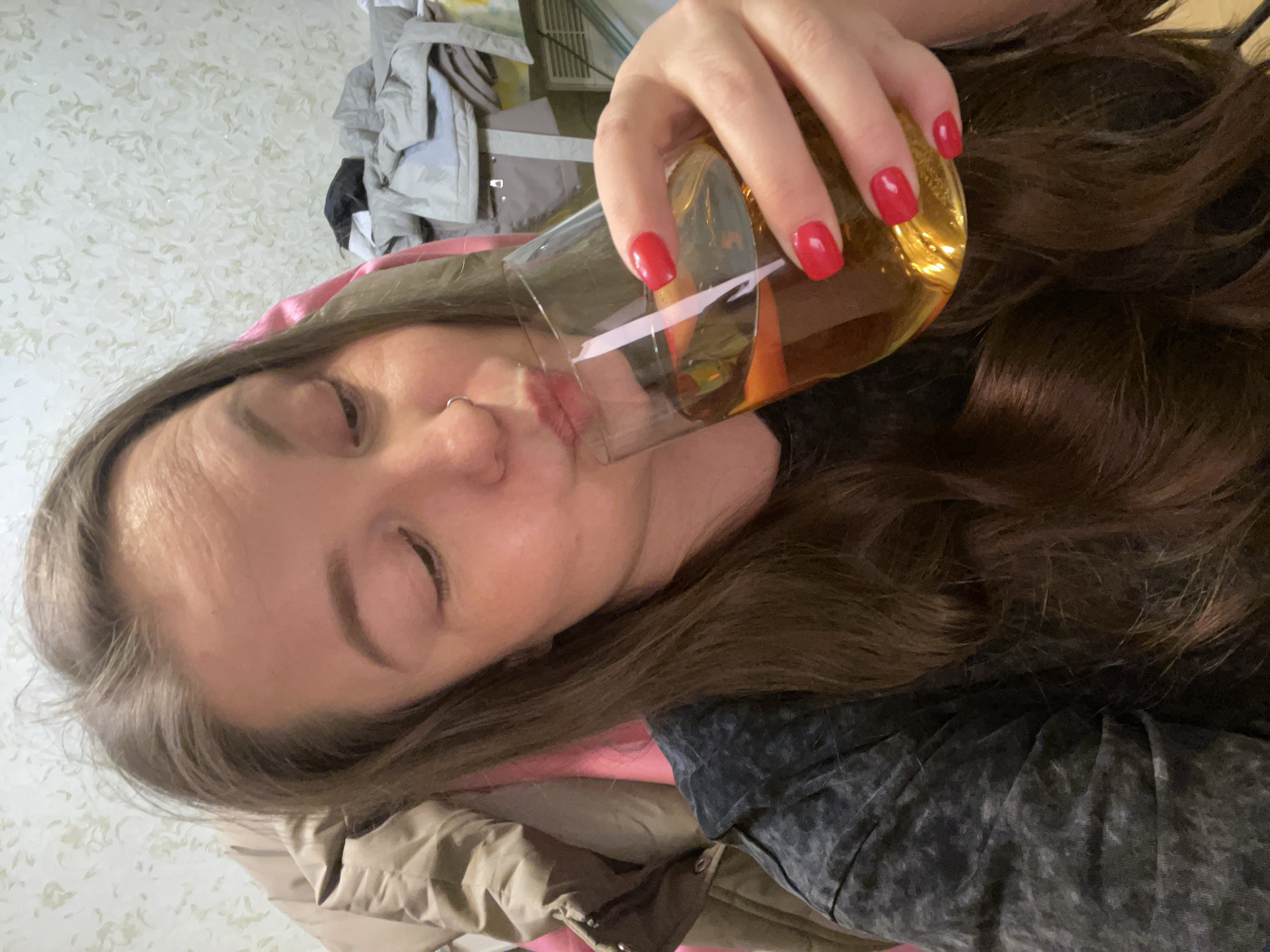 A woman with brown hair and red manicured nails is holding a glass of a light amber liquid to her lips. She has a nose ring and her eyes are closed. She is wearing a gray shirt. Behind her is a pile of clothing. The location cannot be determined.