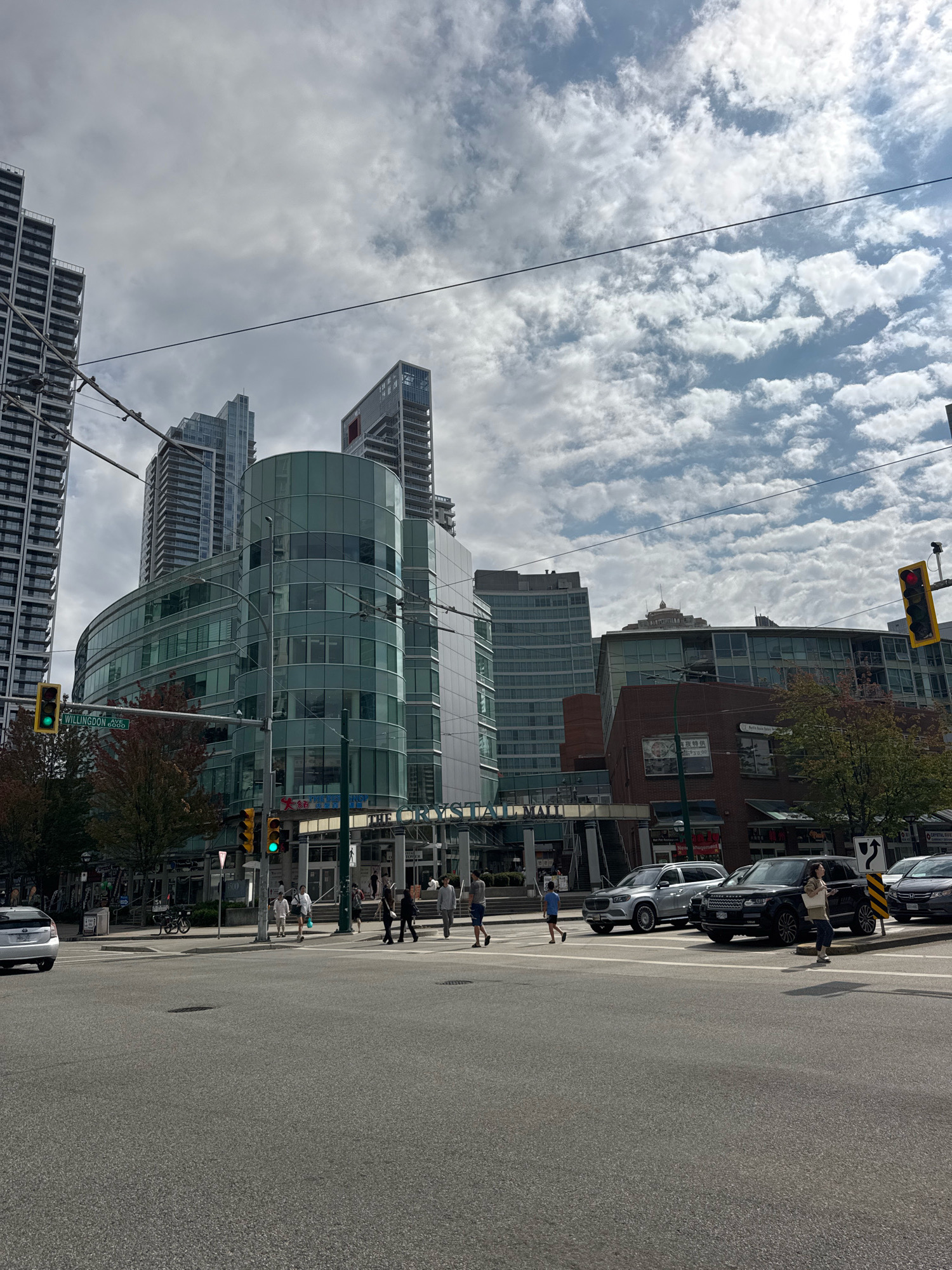 This image depicts an outdoor urban scene with several modern buildings, including high-rise residential towers and a curved glass building housing "THE CRYSTAL" mall. The location is identified as the intersection of Willingdon Avenue and Douglas Place in Burnaby, Canada, indicated by a street sign.
In the foreground, a four-lane asphalt road is visible, with lane markings and a pedestrian crosswalk. Several cars are present: a white Toyota Prius on the left, a silver SUV and a black Range Rover in the middle, and a dark sedan on the right. Pedestrians are crossing the street at the intersection.
The midground features the aforementioned buildings, including "THE CRYSTAL" mall with its entrance. Traffic lights are visible, with one showing green and another red. Trees with some fall foliage are also present.
The background consists of a cloudy sky with dramatic cloud formations. Power lines are strung across the sky, contributing to the urban atmosphere. The overall impression is a bustling city intersection on an overcast day.
