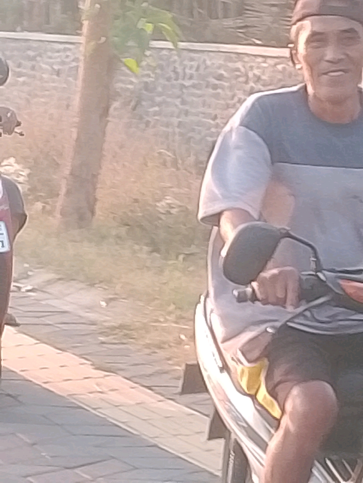 The image depicts an outdoor scene in Probolinggo, Indonesia, featuring a man riding a motorcycle. The man, wearing a cap and a two-toned t-shirt, is partially visible. He appears to be smiling. He is operating the motorcycle, which has a side mirror. The background shows a stone wall and vegetation. The foreground includes a paved walkway. The lighting suggests it is daytime, possibly late afternoon due to the warm tones.
