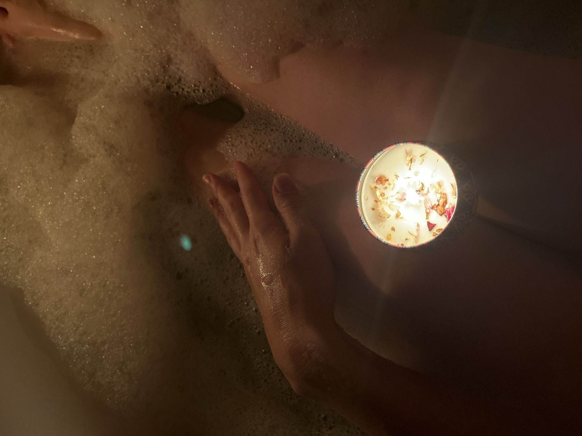 A first-person perspective photo shows a person in a bathtub filled with bubbly water. The person's hand is visible, partially submerged in the water next to a lit candle. The candle sits on a surface next to the tub and casts a soft, warm glow. The location is not discernible.