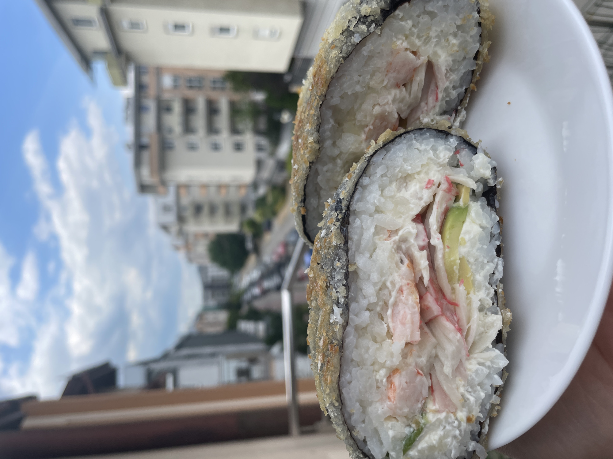A close-up, eye-level, vertical outdoor shot shows a white plate with two halves of a deep-fried sushi roll. The sushi roll is thick with a crispy golden-brown exterior and a visible white rice layer inside. The filling consists of what appears to be imitation crab meat, avocado slices, and a creamy white sauce. The plate is held by a hand, visible at the bottom left. In the blurred background, a city street with buildings and trees can be seen under a cloudy blue sky, suggesting an urban setting. The balcony railing is also visible in the mid-ground.