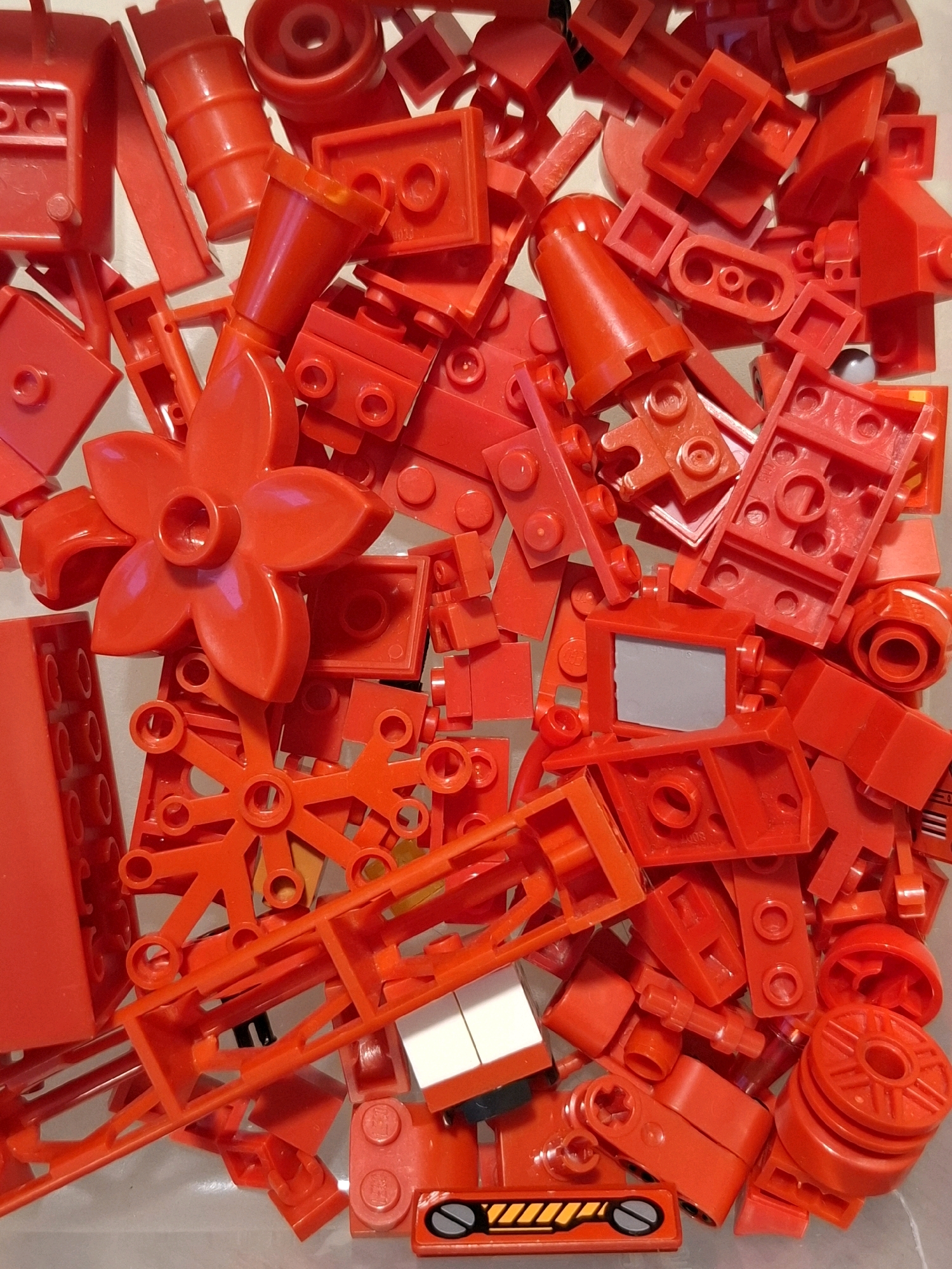 The image displays a close-up, overhead view of a large collection of red plastic building toy pieces, reminiscent of LEGOs. The pieces are scattered and piled together, creating a textured surface. Various shapes and sizes are visible, including bricks, plates, wheels, and decorative elements like a flower. Some pieces are stacked or interlocked, while others lie loose. The lighting is bright, casting subtle shadows that highlight the three-dimensional nature of the toys. The background is a plain, light color, suggesting the toys are contained within a box or on a surface. There is no indication of the city of Tyumen, Russia, in the image.